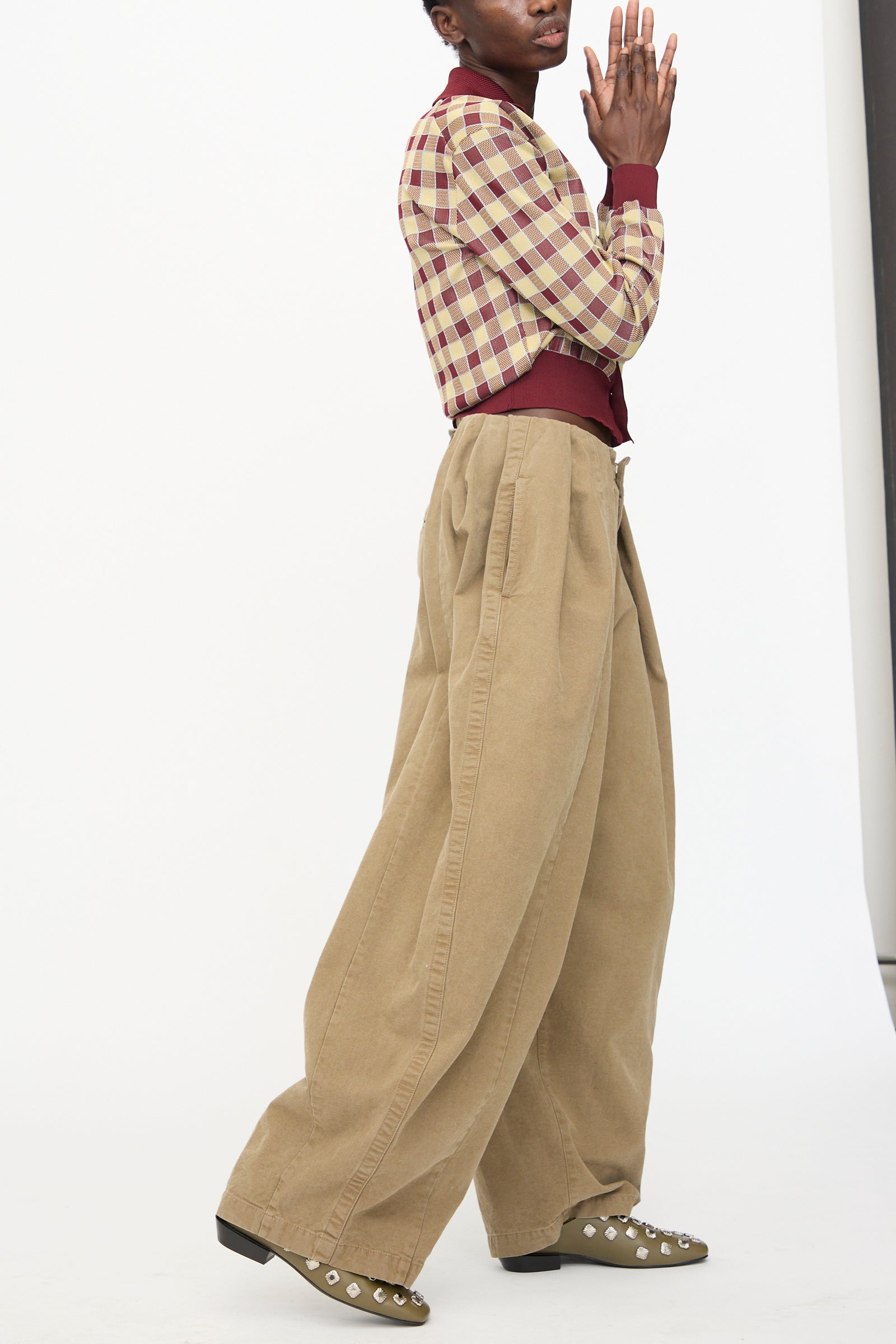 A person poses in TOGA ARCHIVES’ Oxford Wide Pants in Beige with a cotton canvas texture, paired with a plaid jacket and studded olive green shoes against a plain white background.