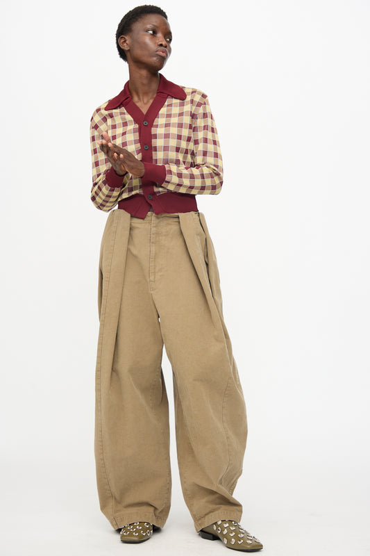 A person stands against a white background, wearing a burgundy and beige checkered shirt, TOGA ARCHIVES Oxford Wide Pants in Beige, and studded khaki shoes, with hands clasped.