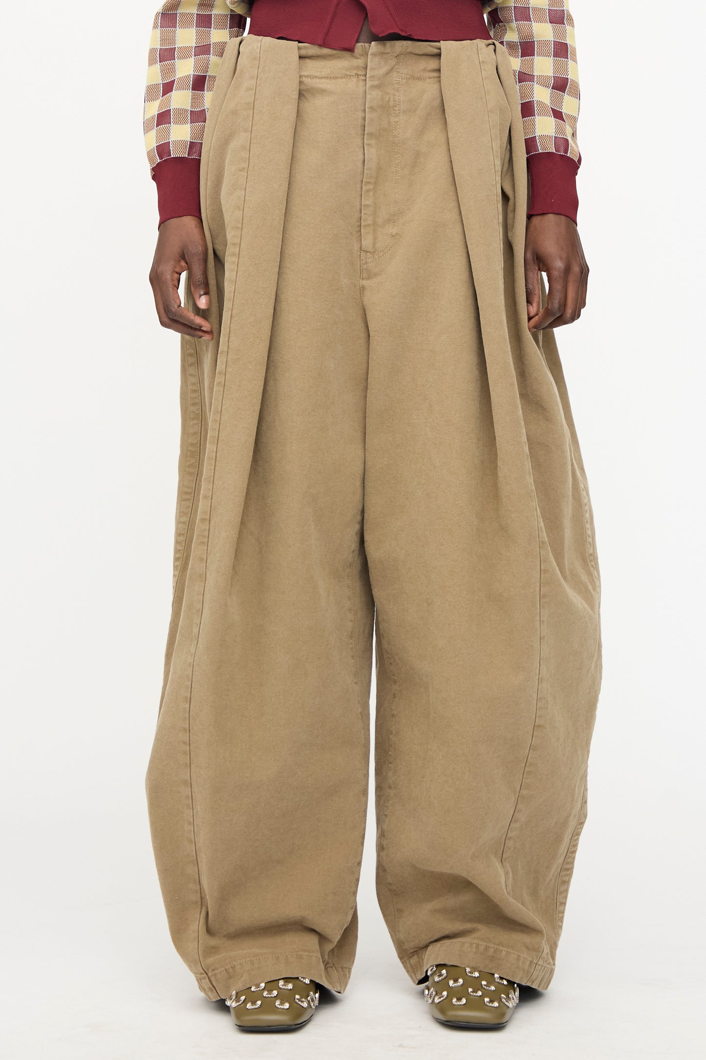 A person wearing TOGA ARCHIVES Oxford Wide Pants in Beige, a plaid long-sleeve shirt, and green shoes with silver studs stands against a plain white background.