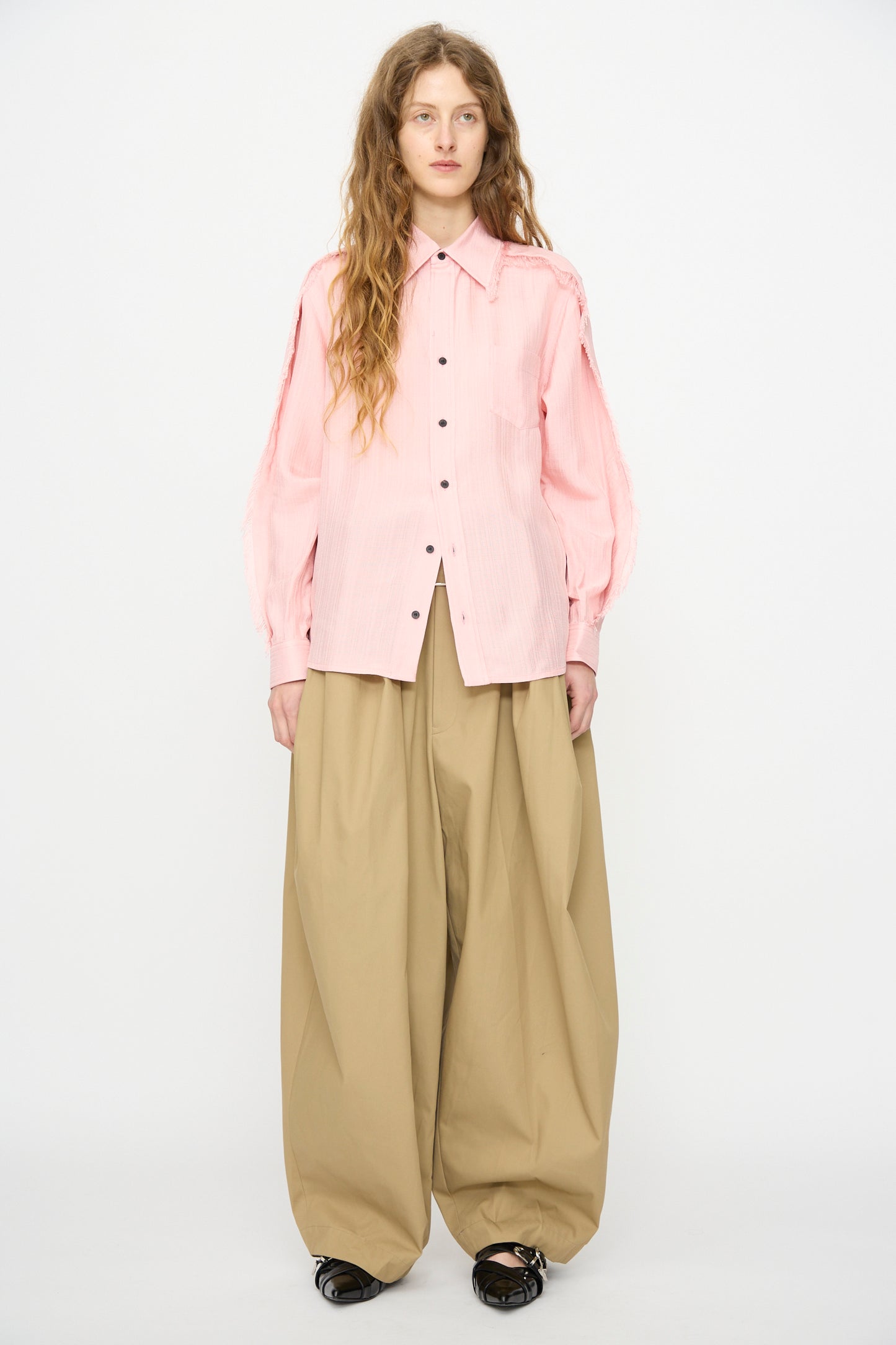 A person stands against a white background wearing the TOGA ARCHIVES Plain Weave Shirt in Pink, wide beige trousers, and black shoes.