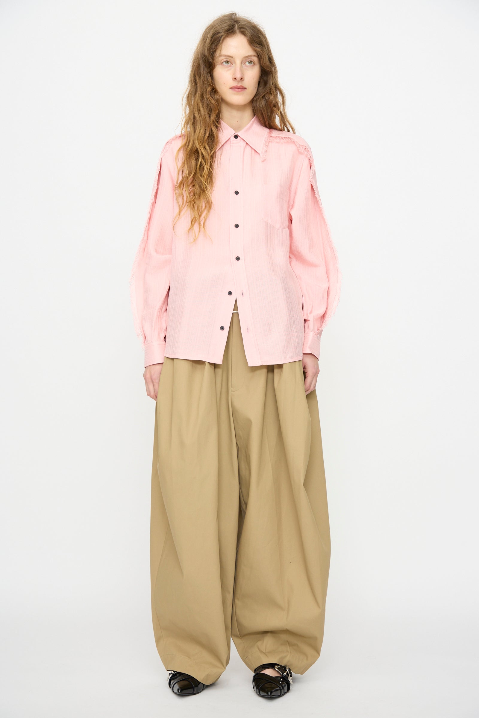 A person stands against a white background wearing the TOGA ARCHIVES Plain Weave Shirt in Pink, wide beige trousers, and black shoes.