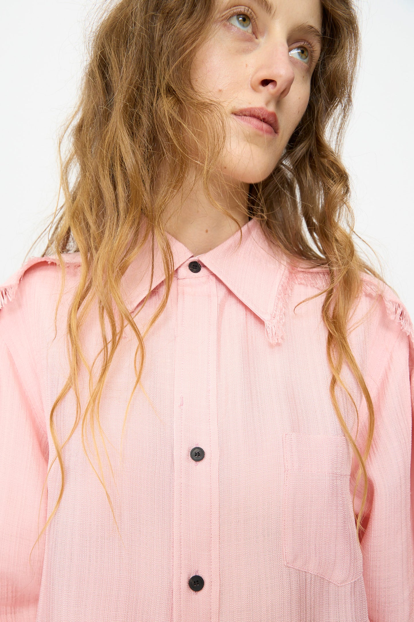 A woman with wavy, light brown hair wears the TOGA ARCHIVES Plain Weave Shirt in Pink, featuring a lightweight rayon fabric, a pocket, and frayed shoulders as she looks slightly upward.