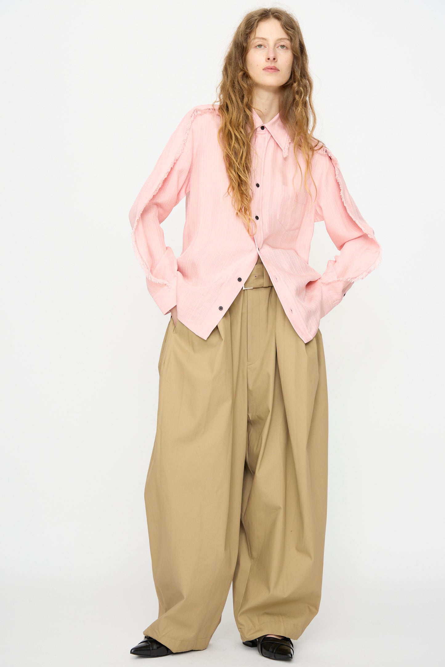 A person wears the TOGA ARCHIVES Plain Weave Shirt in Pink with wide, high-waisted tan pants, standing against a plain white background.