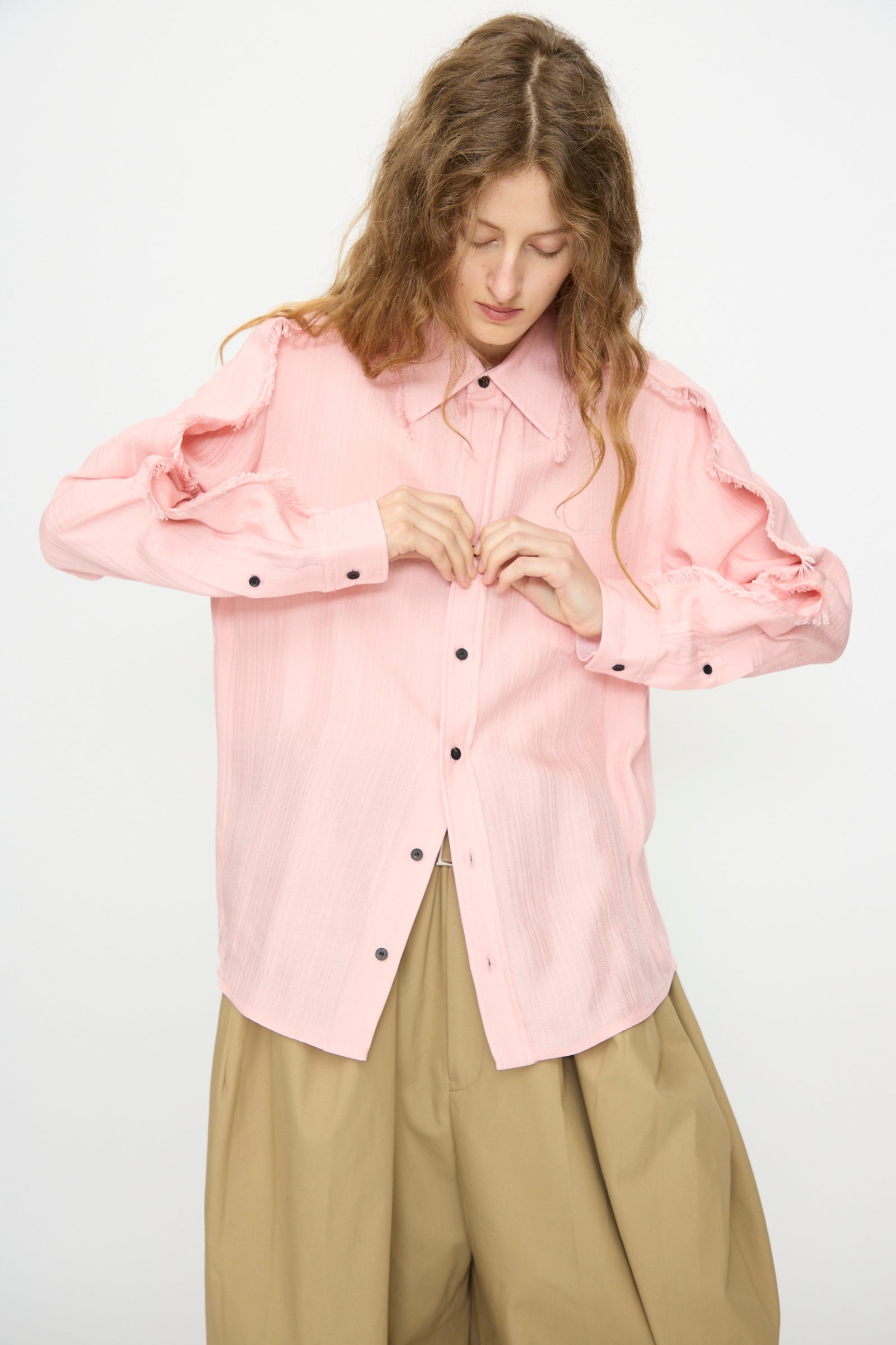 A person with long hair buttons the Plain Weave Shirt in Pink by TOGA ARCHIVES, featuring frayed shoulder seams and a western style, paired with loose tan pants against a plain light background.