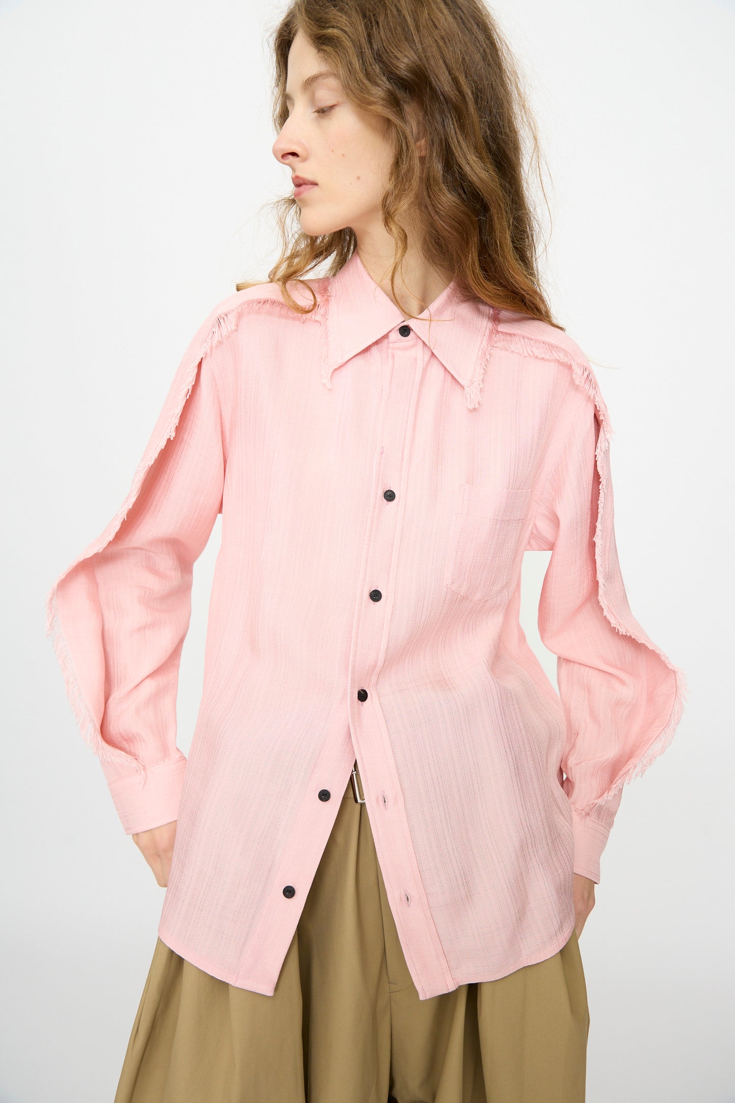A person wears the TOGA ARCHIVES Plain Weave Shirt in Pink, featuring open, fringed sleeve seams, paired with khaki pants against a plain background.