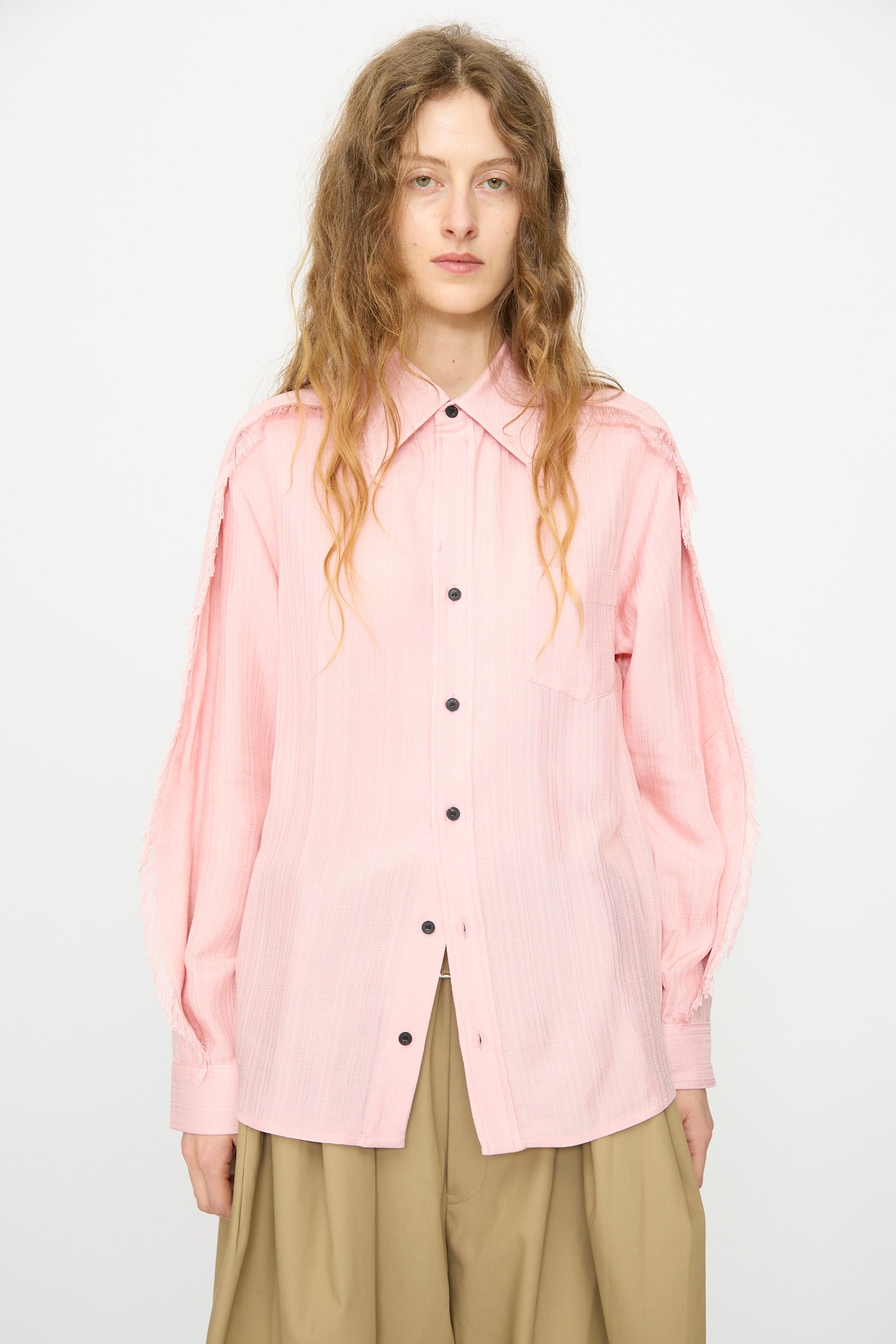 A person with long wavy hair wears the TOGA ARCHIVES Plain Weave Shirt in Pink with black buttons and beige pants, standing against a plain white background.