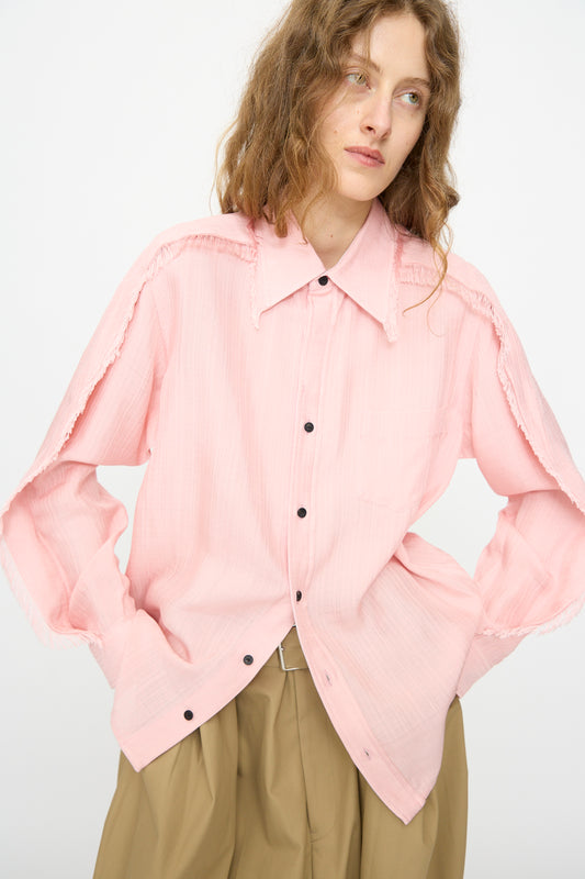 A person with wavy, light brown hair wears the TOGA ARCHIVES Plain Weave Shirt in Pink— a lightweight, western-style button up with frayed shoulder seams—paired with tan high-waisted pants against a plain background.