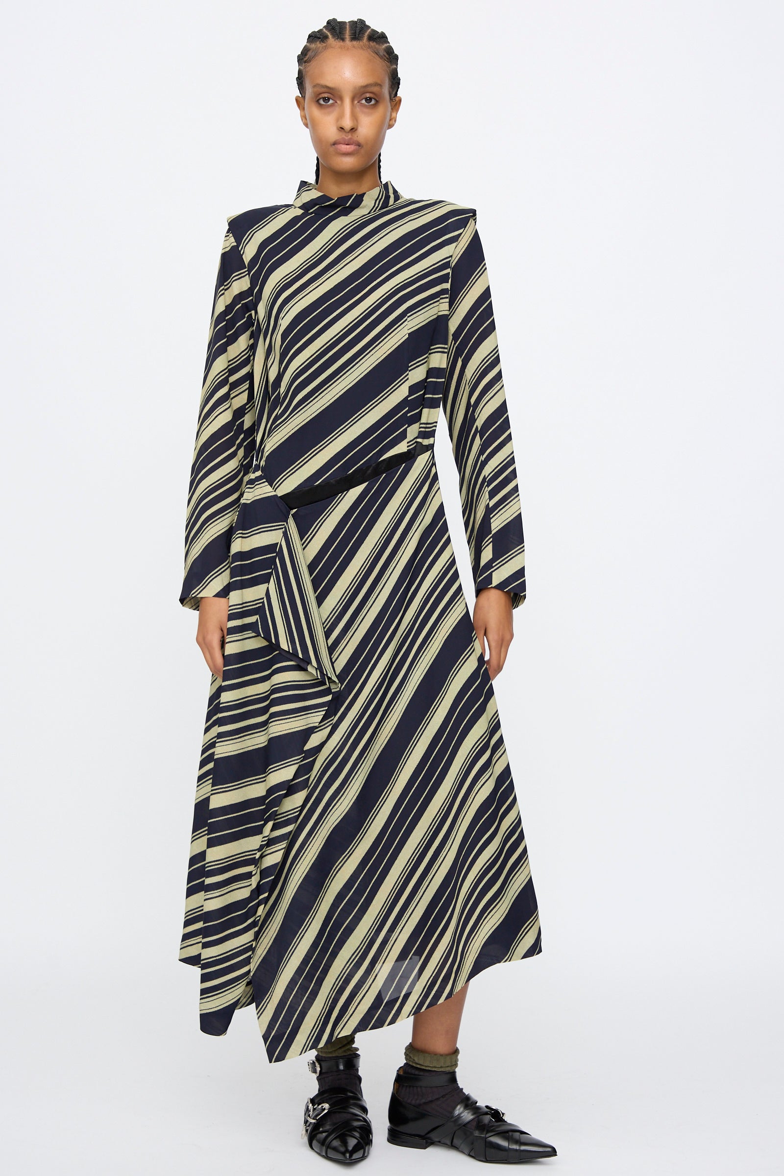 A person stands against a white background wearing the TOGA ARCHIVES Rayon Print Dress in Navy, a long sleeve maxi dress made in Japan with an asymmetrical black design, beige diagonal stripes, and paired with black shoes.