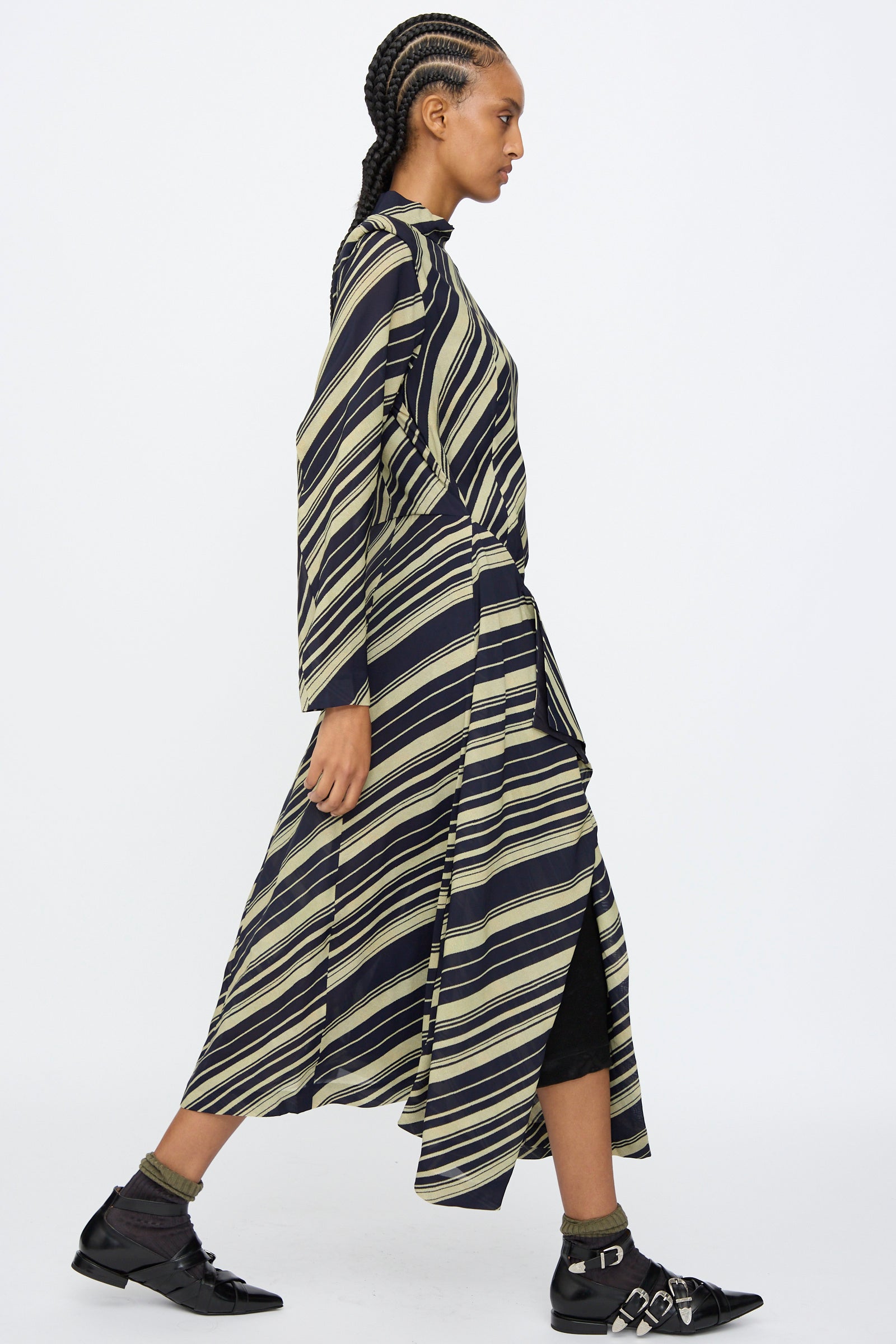 A person in profile wears the TOGA ARCHIVES Rayon Print Dress in Navy, featuring diagonal navy and beige stripes. The long sleeve maxi dress is made in Japan and styled with black shoes, socks, and braided hair.