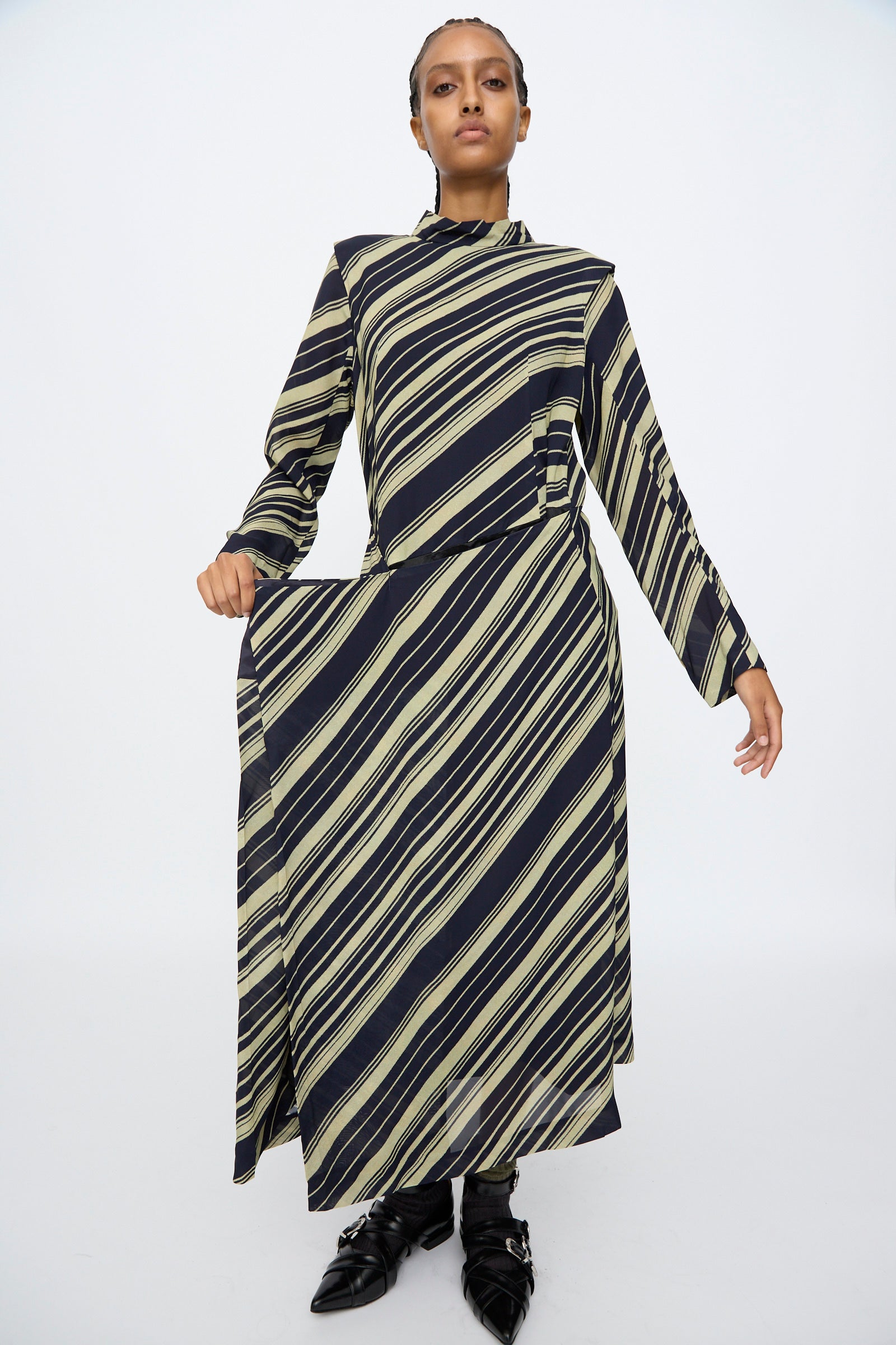 A person stands facing forward, wearing the TOGA ARCHIVES Rayon Print Dress in Navy with long sleeves and diagonal black and beige stripes, paired with black pointed shoes. This elegant dress is made in Japan.