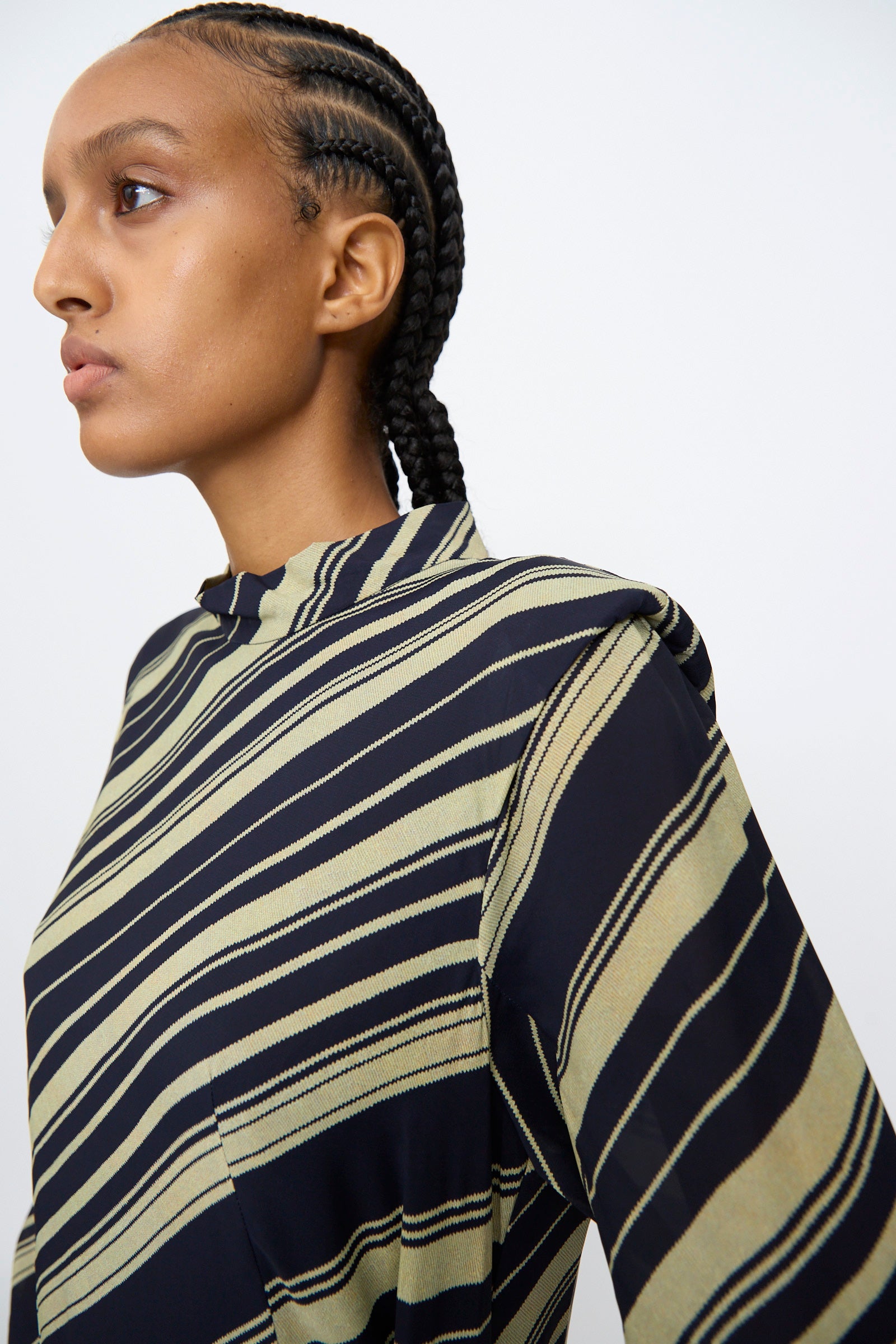 A person with braided hair poses against a plain light background, wearing the TOGA ARCHIVES Rayon Print Dress in Navy, a long sleeve maxi dress with diagonal black and beige stripes.