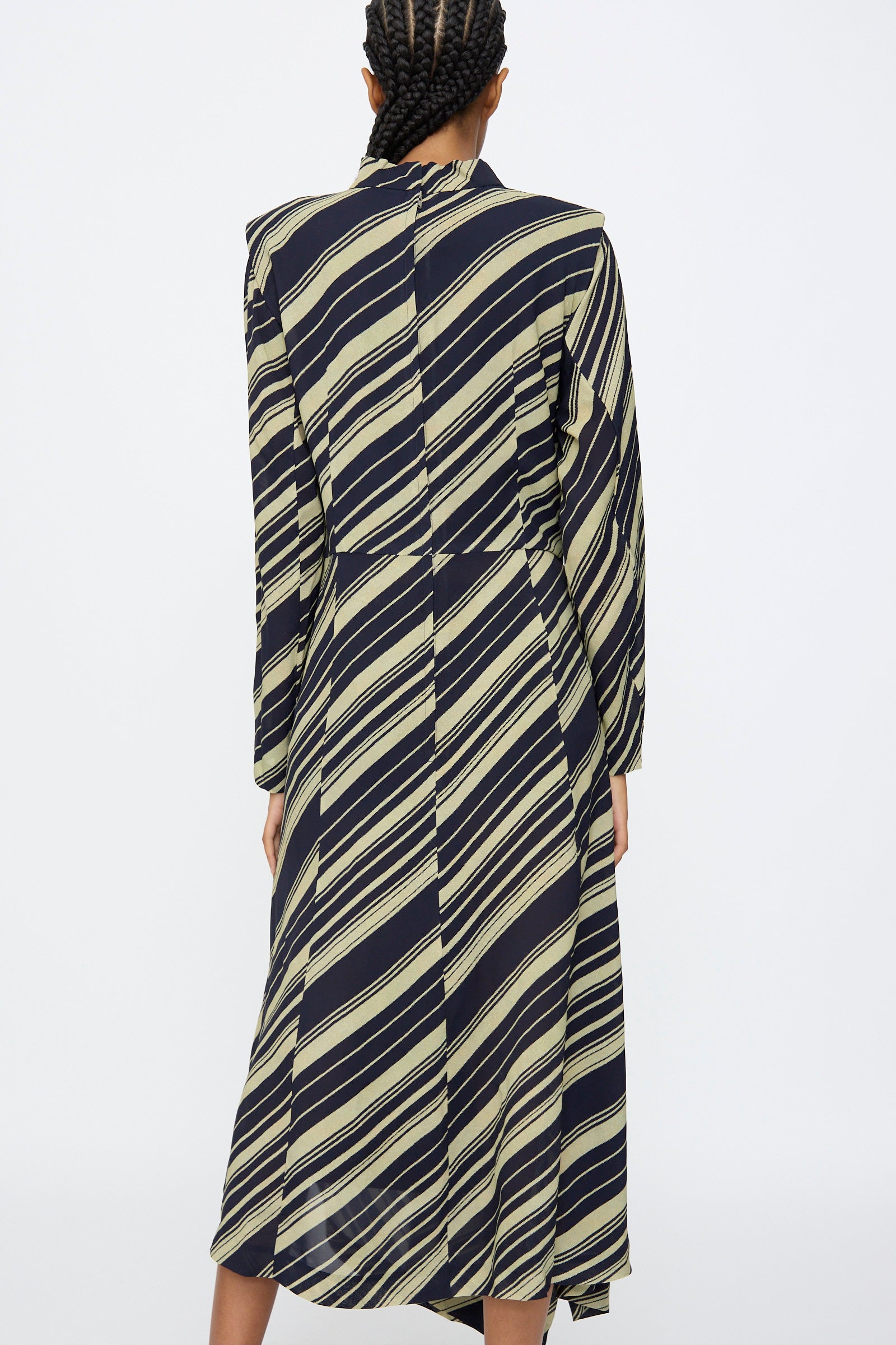 A person with braided hair is shown from the back wearing the TOGA ARCHIVES Rayon Print Dress in Navy, a long sleeve maxi dress made in Japan with diagonal beige stripes on black fabric.