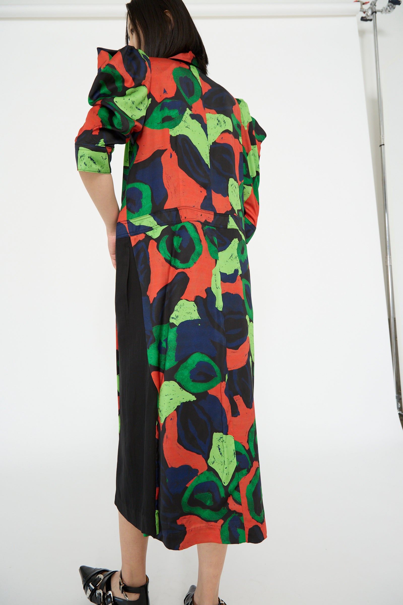 A person in a TOGA ARCHIVES Satin Print Dress in Mix with long, colorful puff sleeves and a bold red, green, and black abstract pattern stands facing away.