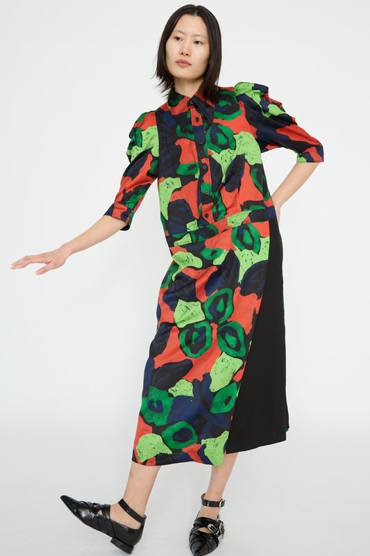 A model wears the TOGA ARCHIVES' Satin Print Dress in Mix with abstract floral designs in red, green, and black, paired with black shoes, on hand on hip against a white backdrop. 