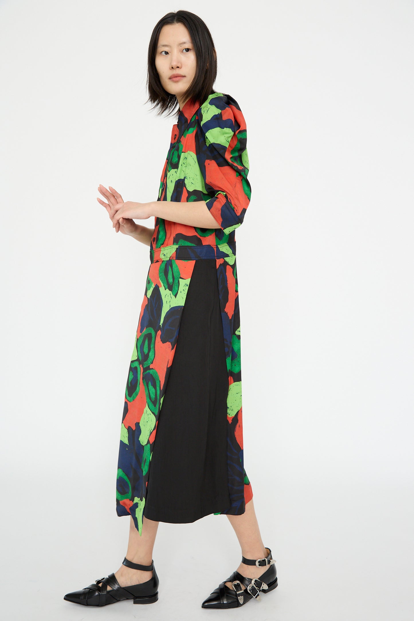 A model poses against a plain white background, wearing the TOGA ARCHIVES Satin Print Dress in Mix, with colorful abstract patterns and puff sleeves, paired with black shoes.