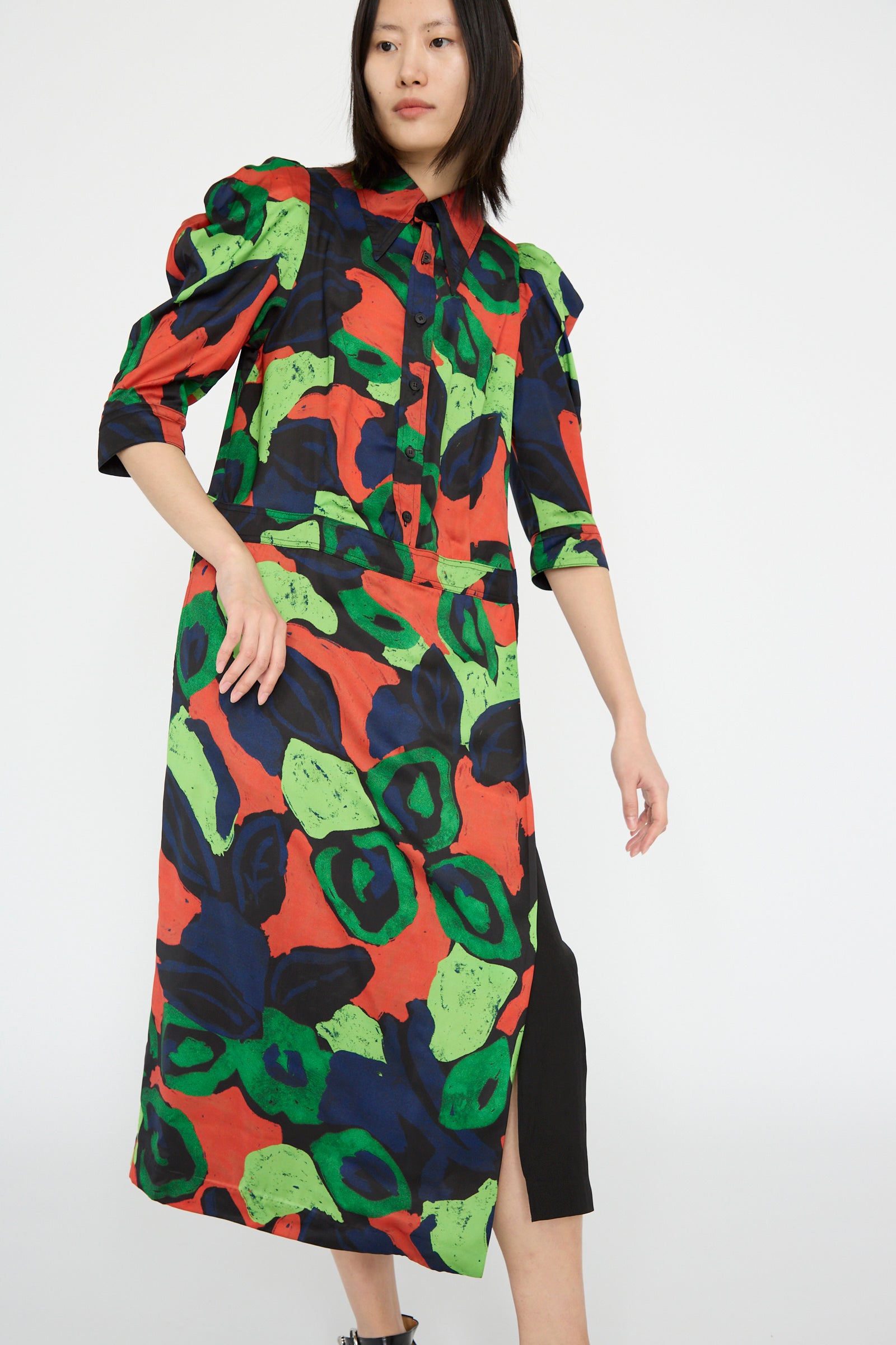 A person is dressed in the Satin Print Dress in Mix by TOGA ARCHIVES, with a bold abstract pattern in red, green, and blue with 3/4 puff-like sleeves and a slit.