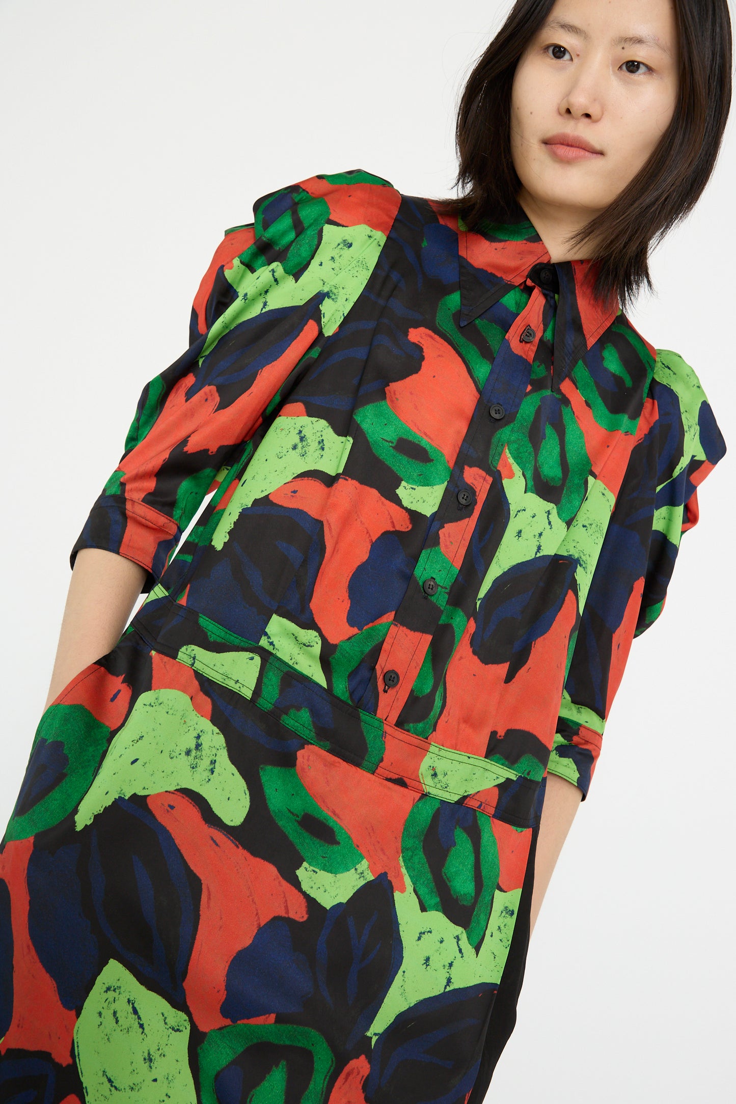 A person wearing the TOGA ARCHIVES Satin Print Dress in Mix, featuring colorful abstract patterns with green, red, and black shapes and a drop waist, against a white backdrop.