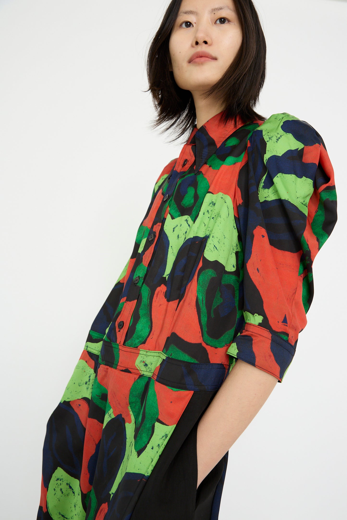 Wearing a TOGA ARCHIVES Satin Print Dress in Mix, featuring colorful abstract patterns with red, green, and blue hues, a person looks toward the camera against a plain background.