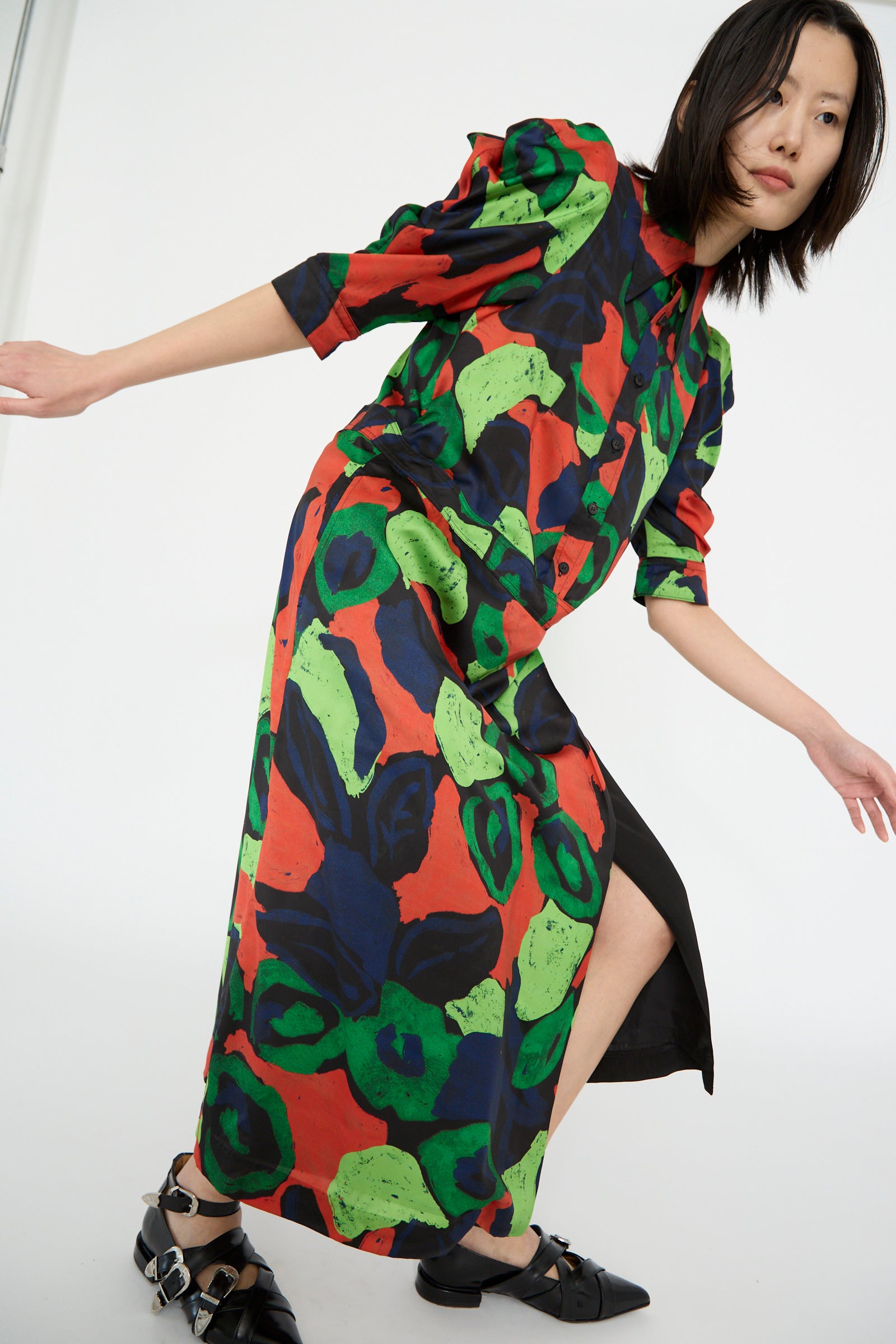 A model striking a dynamic pose in a TOGA ARCHIVES Satin Print Dress in Mix, with black shoes, against a plain backdrop.