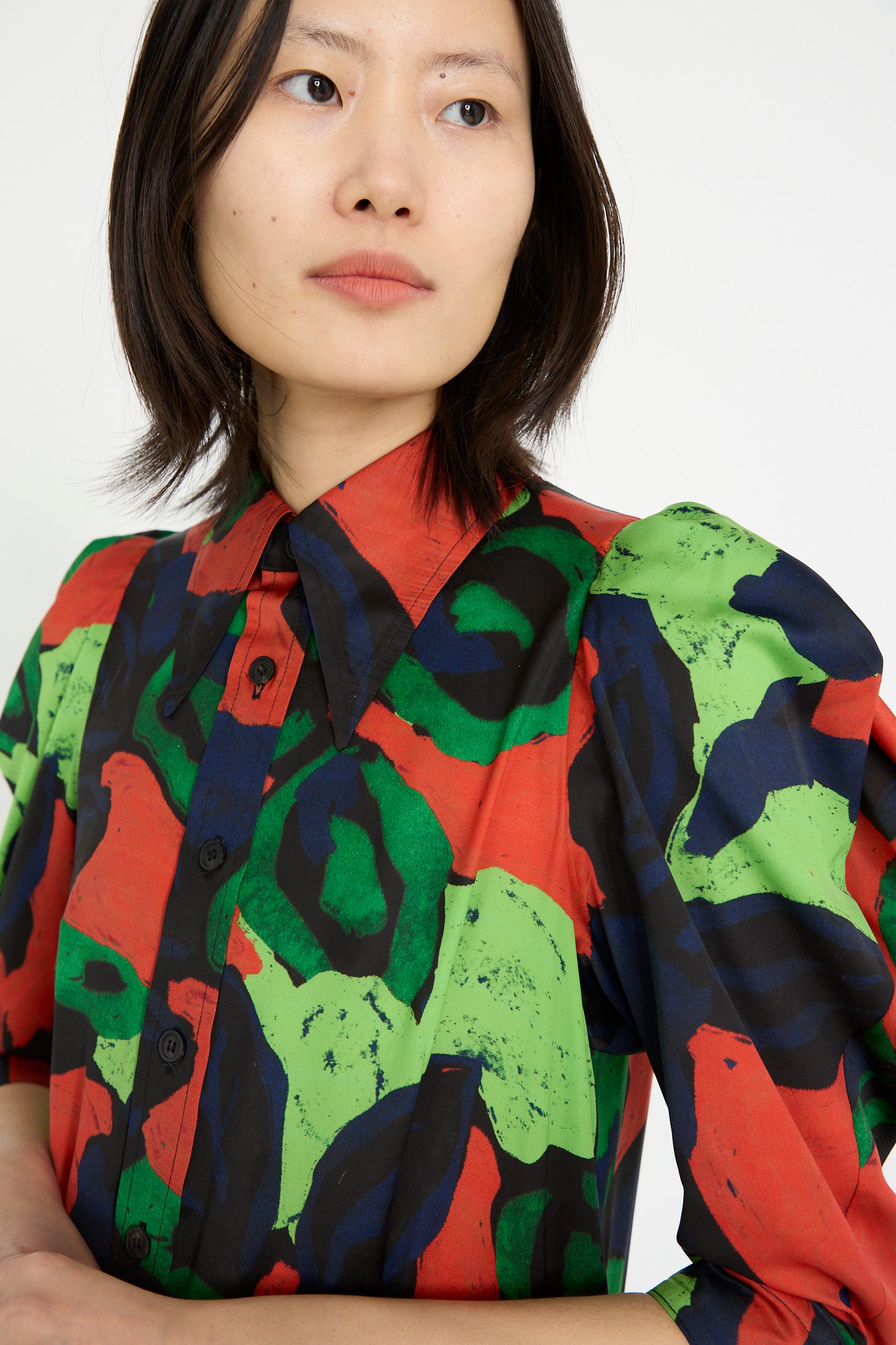 A model in a TOGA ARCHIVES Satin Print Dress in Mix, featuring vibrant red, green, and black abstract patterns, gazes sideways against a white backdrop.