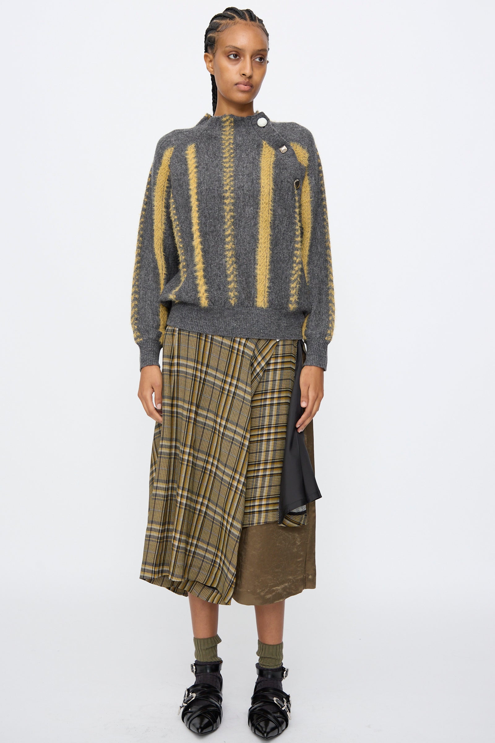 A person stands against a plain background wearing the TOGA ARCHIVES Stripe Knit Pullover in Gray, styled with a layered plaid and solid skirt, plus black shoes and socks.
