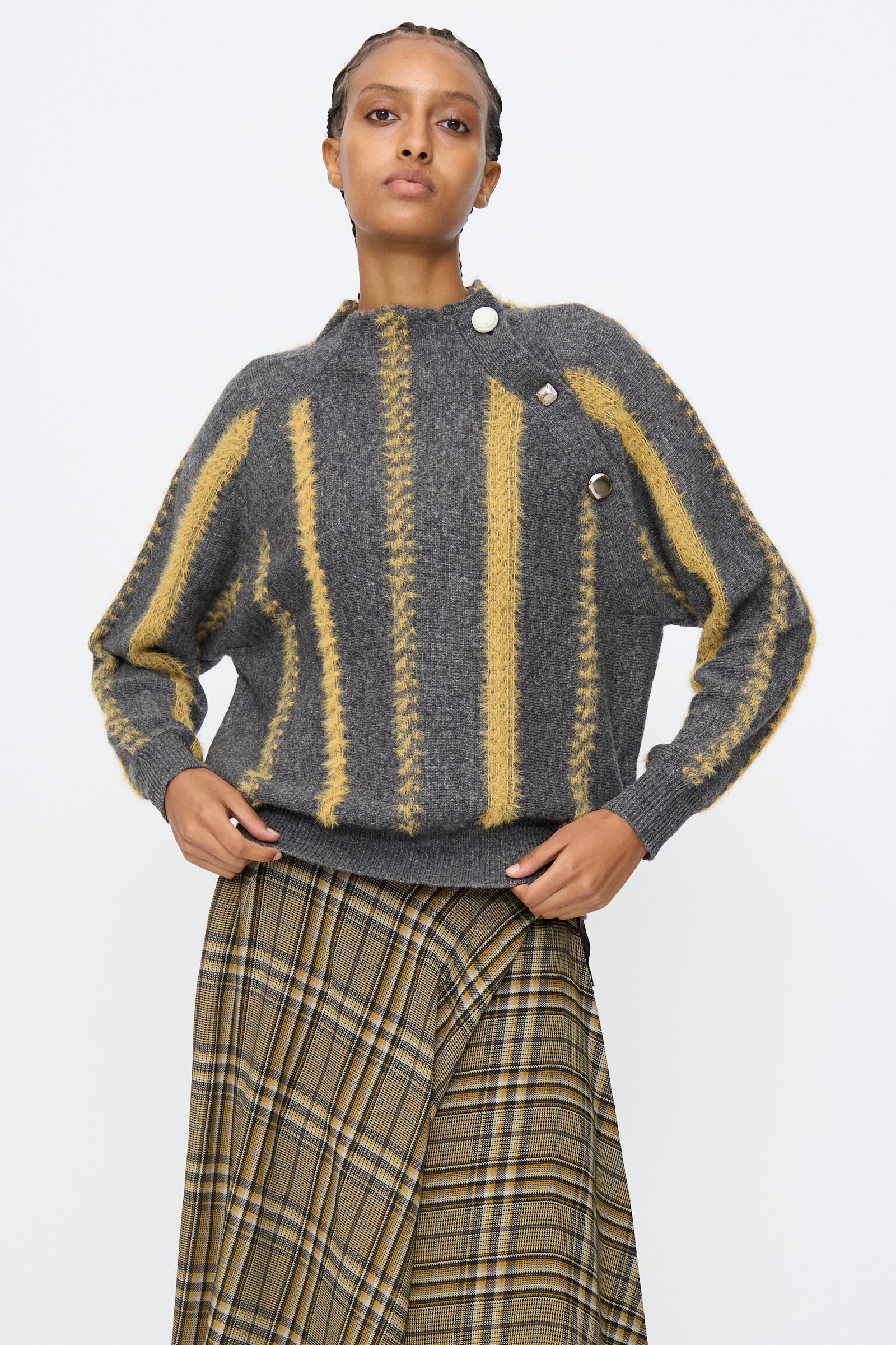 A person wears the TOGA ARCHIVES Stripe Knit Pullover in Gray, featuring yellow vertical stripes and buttons, paired with a brown and yellow plaid skirt, standing against a plain white background.