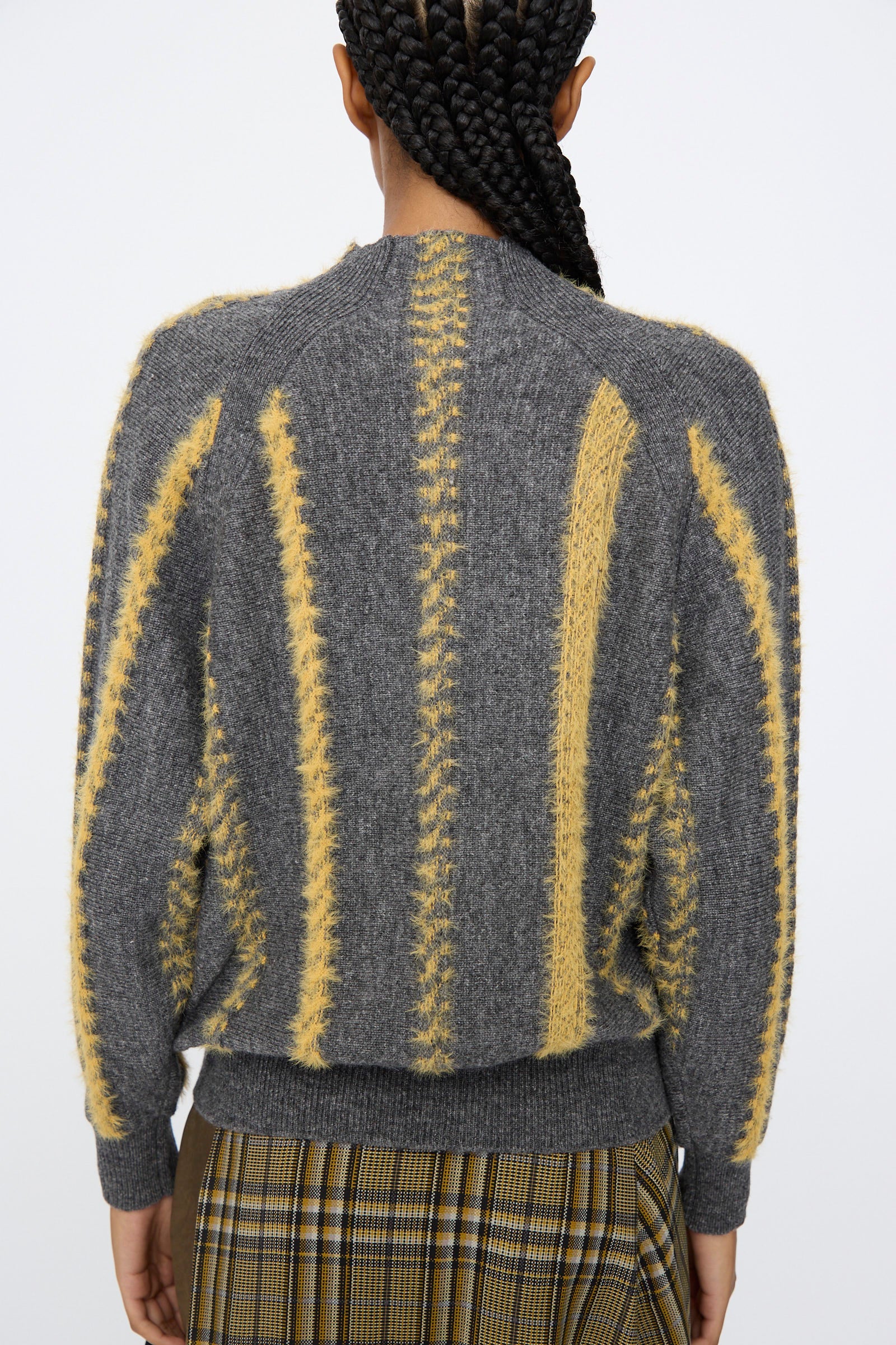 A person with braided hair is seen from the back, wearing the TOGA ARCHIVES Stripe Knit Pullover in Gray, featuring yellow textured vertical stripes, paired with a plaid skirt against a plain background.