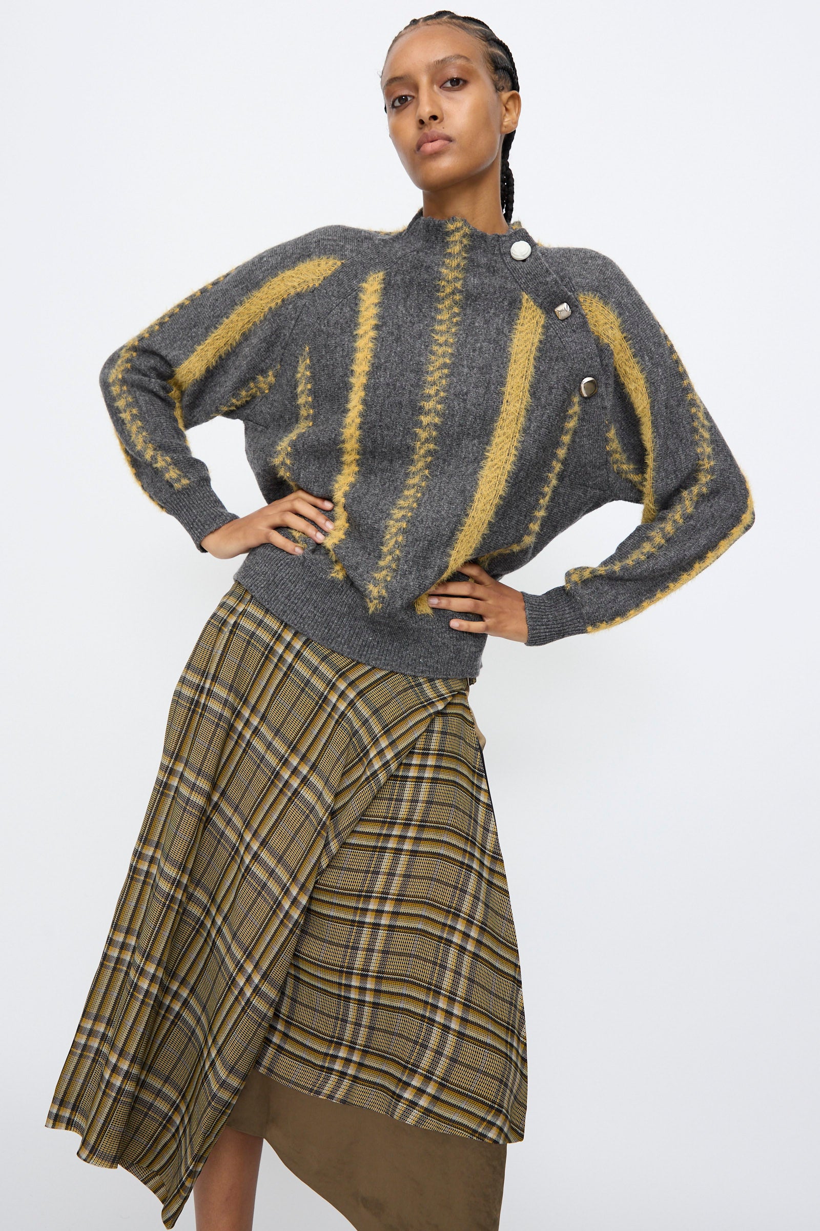 A person models the TOGA ARCHIVES Stripe Knit Pullover in Gray, featuring yellow stripes and button details, paired with an asymmetrical plaid skirt against a plain white background.