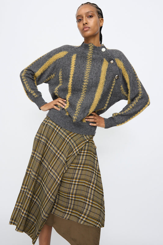A person models the TOGA ARCHIVES Stripe Knit Pullover in Gray, featuring yellow stripes and button details, paired with an asymmetrical plaid skirt against a plain white background.