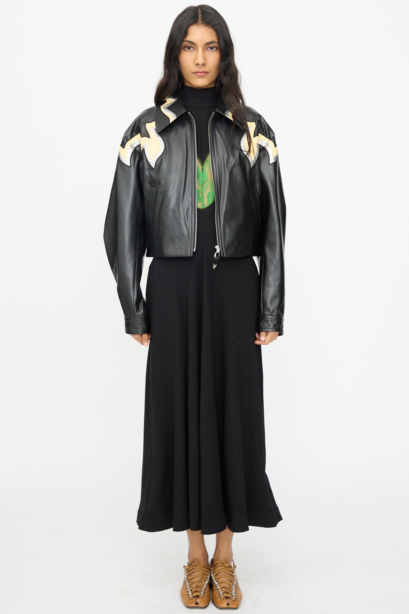 A person faces forward in a black maxi dress and tan woven shoes, wearing the TOGA ARCHIVES Synthetic Leather Jacket in Black with white western motifs on the shoulders, set against a plain white background.