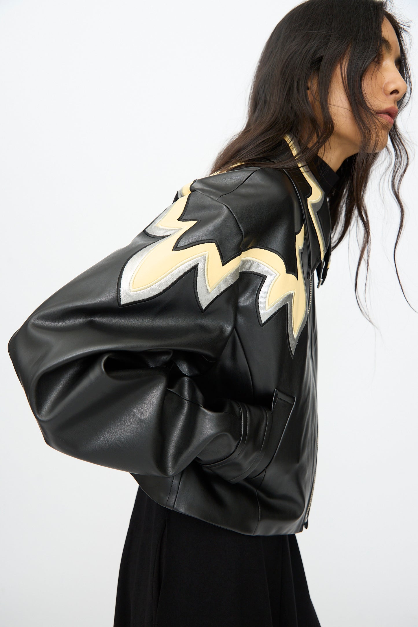 A person with long dark hair wears the TOGA ARCHIVES Synthetic Leather Jacket in Black, featuring beige and white zigzag patterns with a subtle western motif on the shoulders, set against a plain light background.