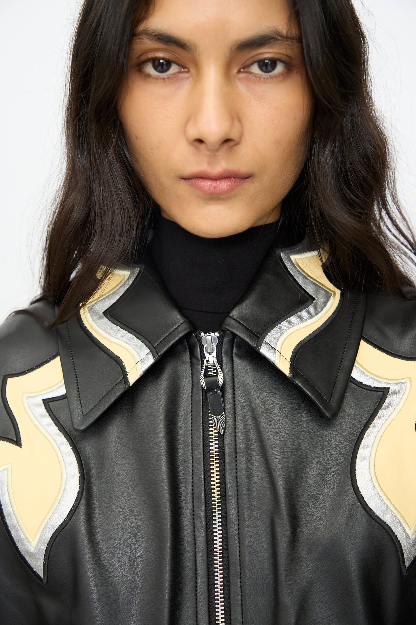 A person with long dark hair wears the TOGA ARCHIVES Synthetic Leather Jacket in Black, featuring wavy cream and silver western motifs on the collar and shoulders, over a black turtleneck.