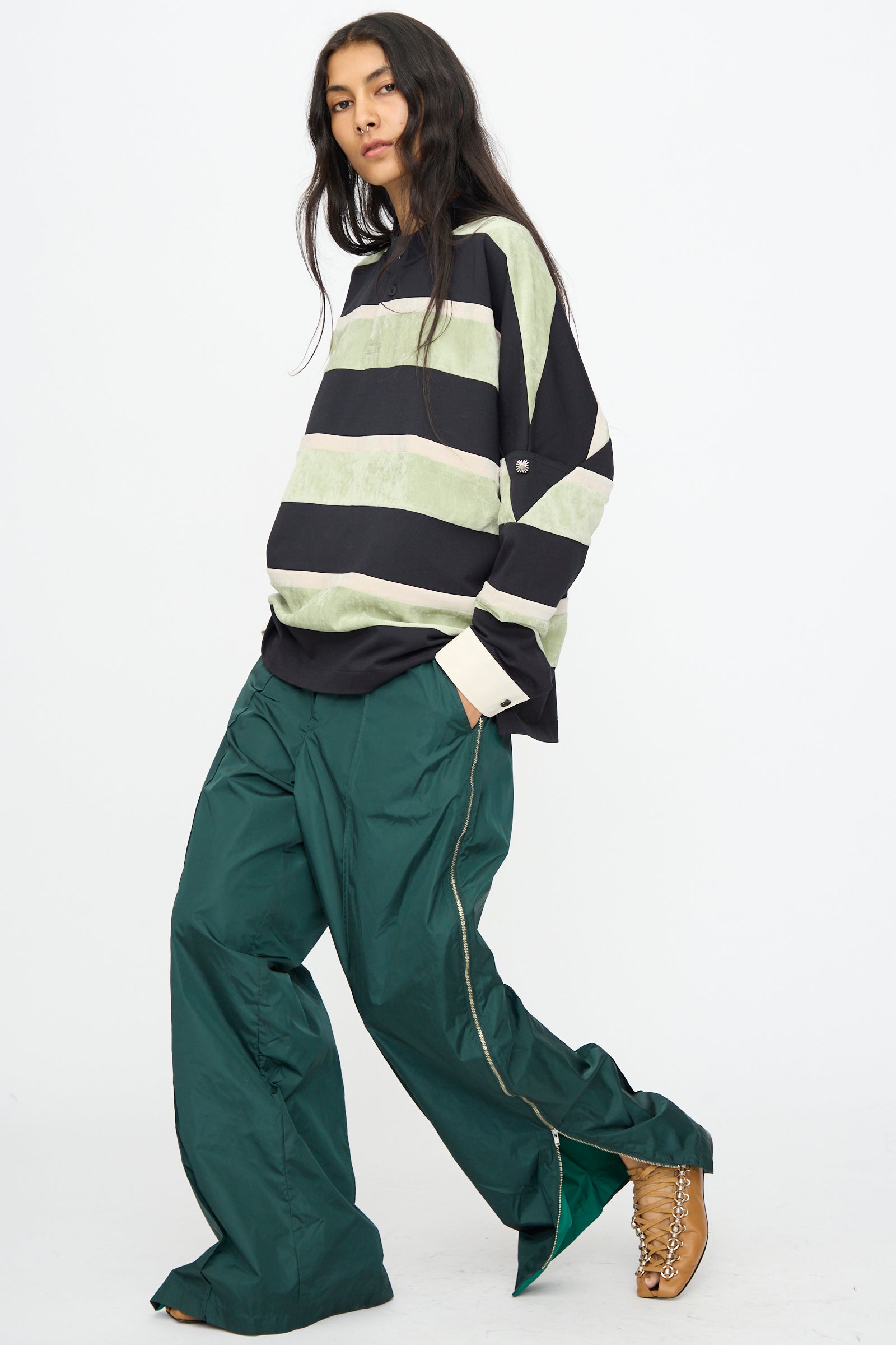 A person wears a loose black and light green striped shirt, TOGA ARCHIVES Taffeta Zip Pant in Green made in Japan, and beige heeled lace-up shoes, standing against a plain white background.