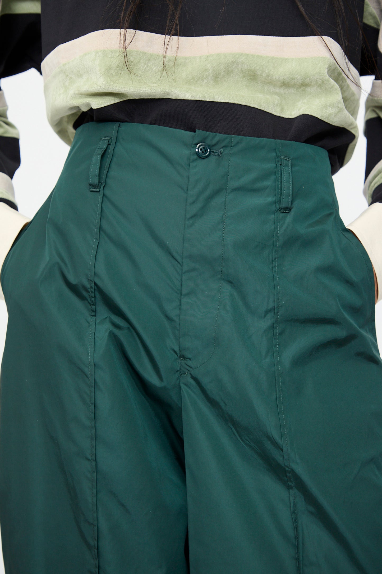 A person wearing TOGA ARCHIVES Taffeta Zip Pant in Green—crafted in Japan—paired with a striped long-sleeve shirt, hands in pockets.