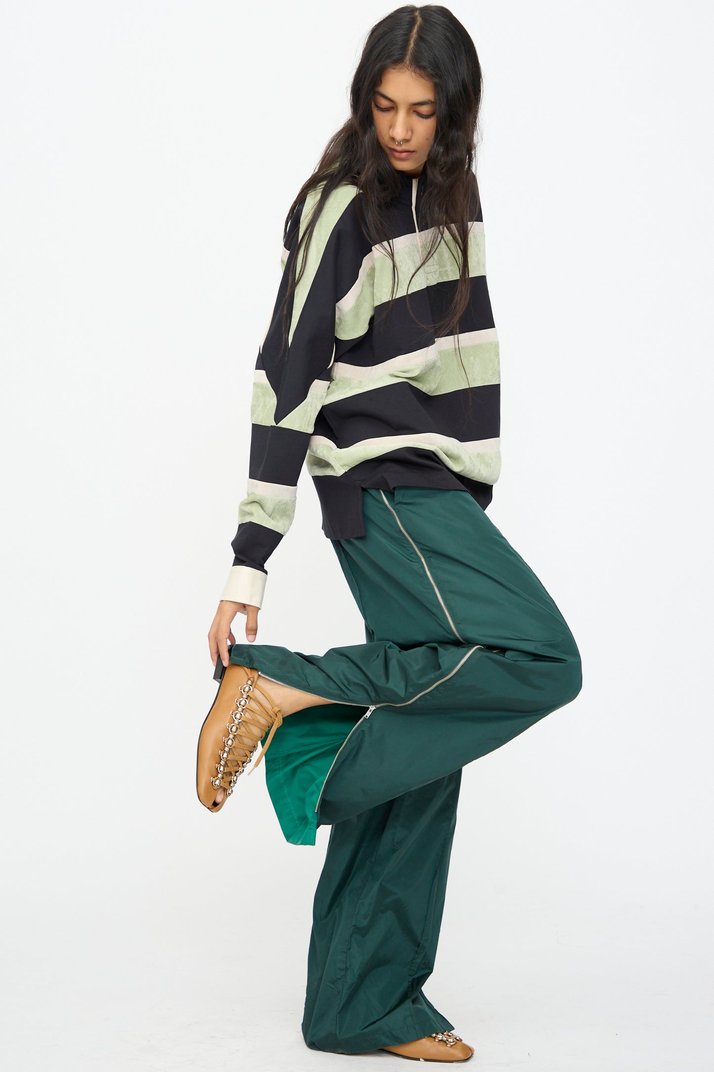 A person poses against a plain white background wearing a black and green striped sweater, TOGA ARCHIVES Taffeta Zip Pant in Green, and tan chain-embellished shoes.