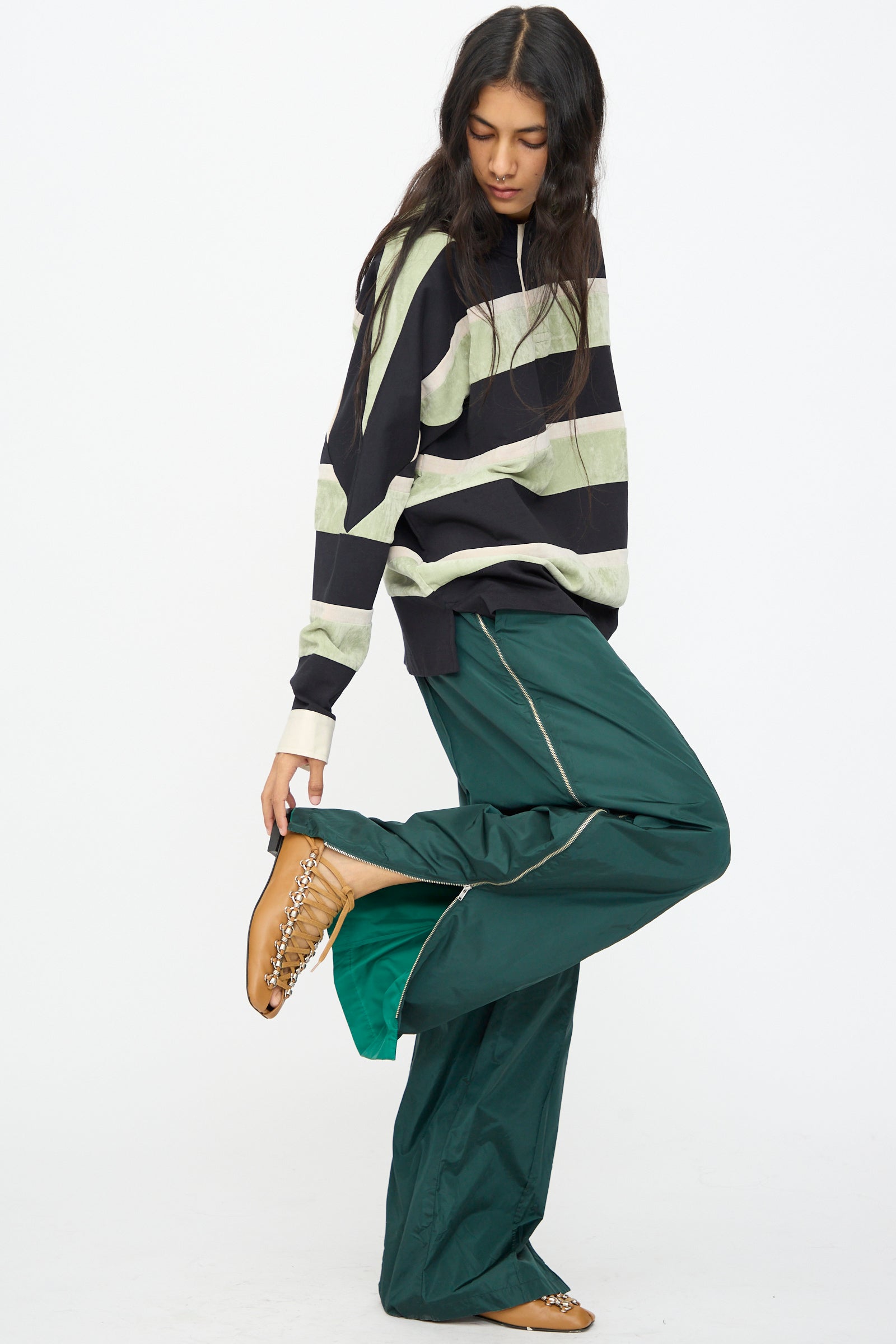 A person poses against a plain white background wearing a black and green striped sweater, TOGA ARCHIVES Taffeta Zip Pant in Green, and tan chain-embellished shoes.