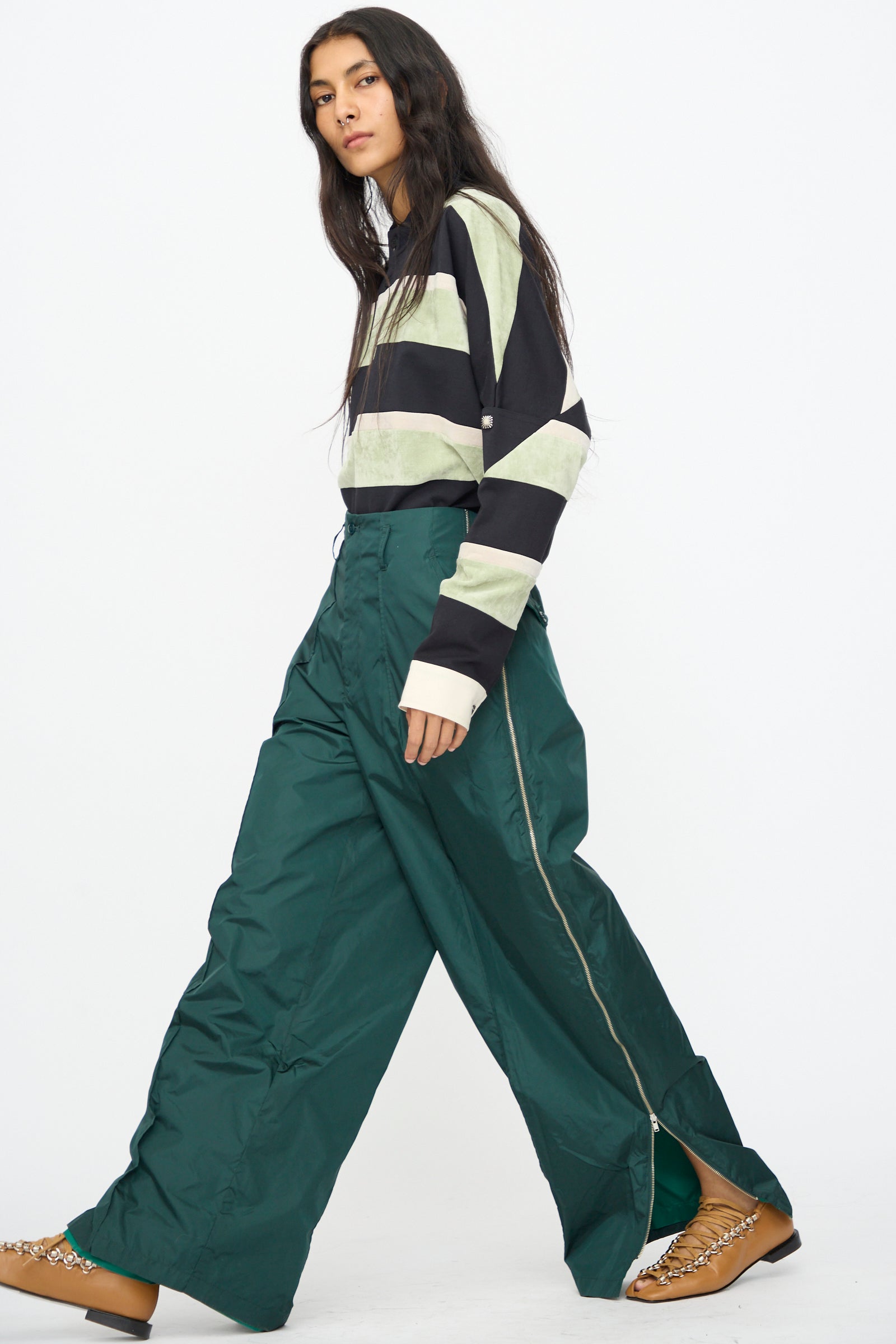 A person with long dark hair wears a black and light green striped top, TOGA ARCHIVES Taffeta Zip Pant in Green, brown shoes, and is walking against a plain white background.
