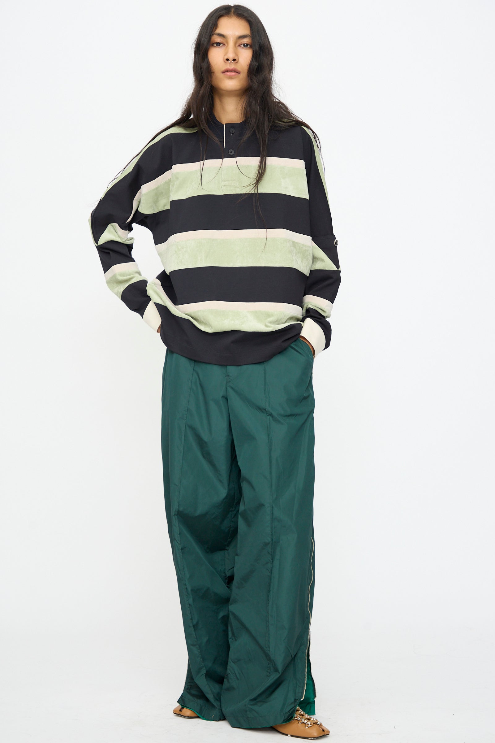 A person with long dark hair wears a loose striped sweatshirt, TOGA ARCHIVES Taffeta Zip Pant in Green made in Japan, and sandals, standing against a plain background.