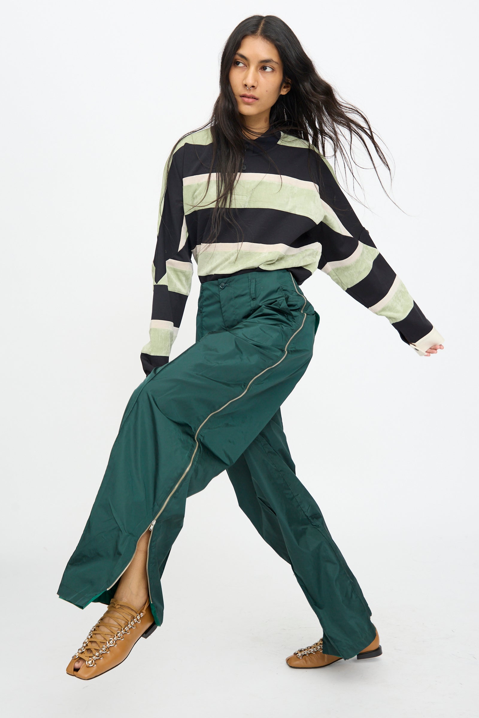 A person wearing a black and light green striped long-sleeve top, TOGA ARCHIVES Taffeta Zip Pant in Green, and tan chain sandals poses against a white background.