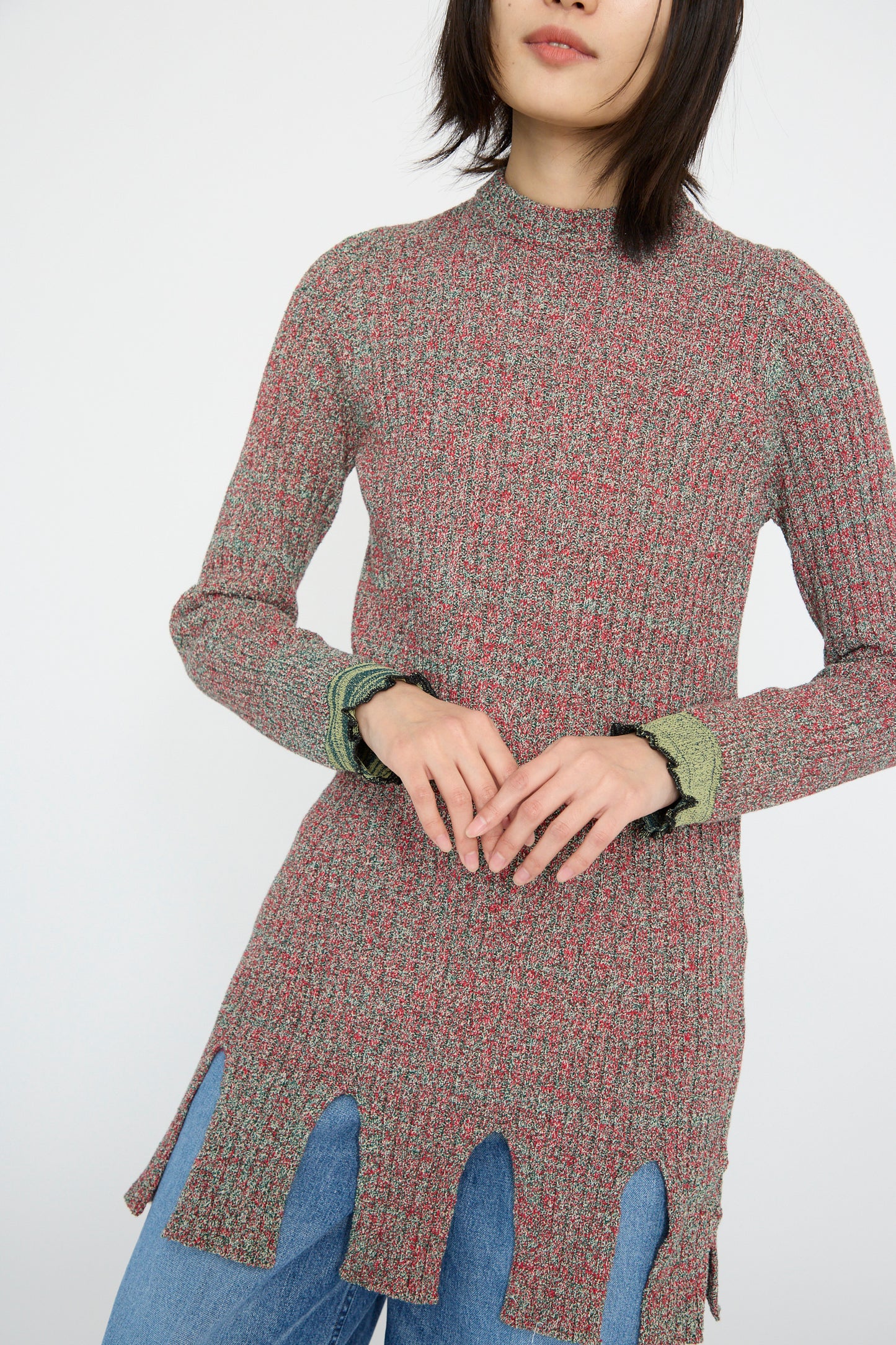 Wearing the TOGA ARCHIVES Tweed Knit Back Zip Pullover in Green with blue jeans, the model showcases a speckled red and green pattern. The outfit, highlighted by a subtle fringe hem and scalloped detail, is set against a plain background.