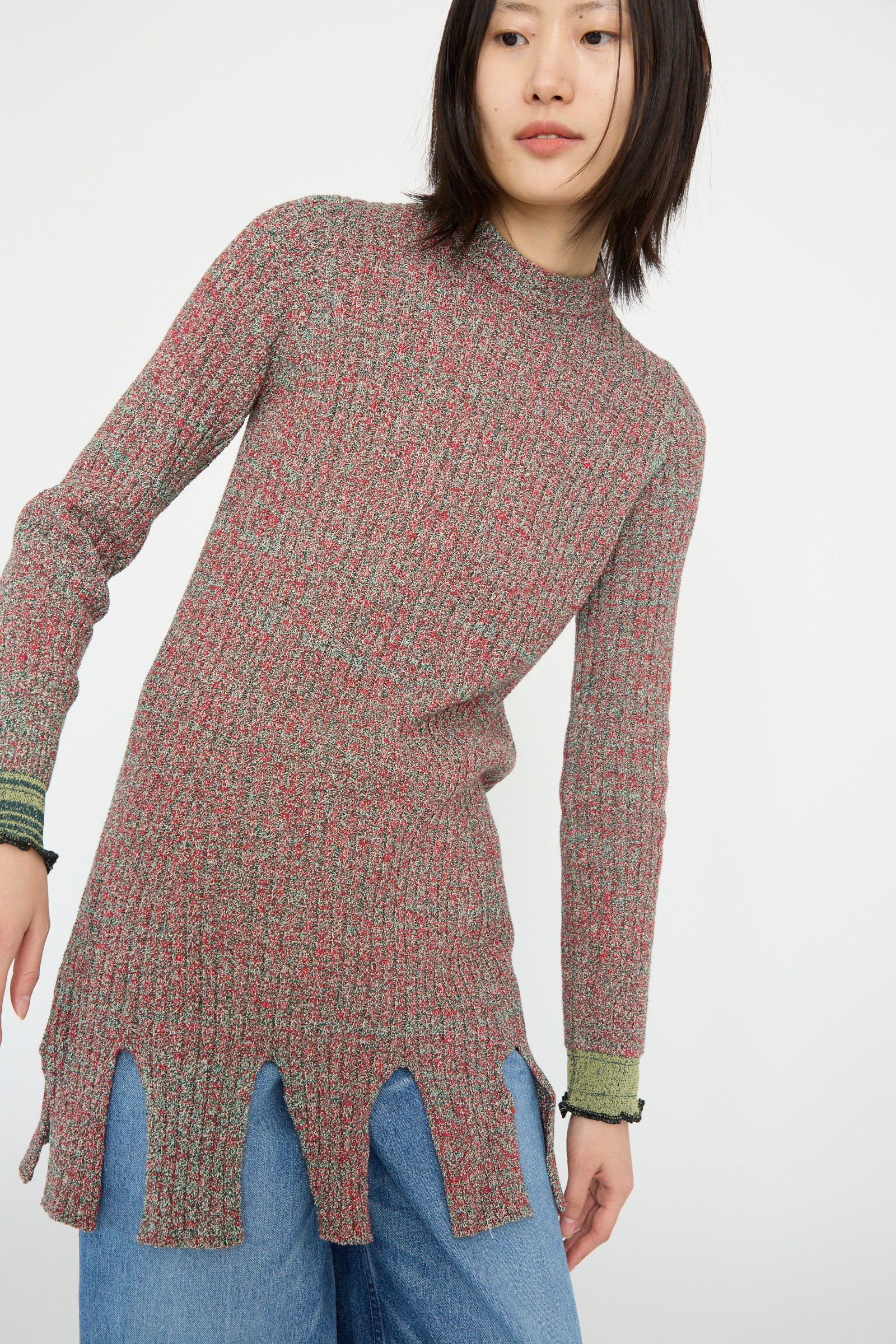 A person wearing a TOGA ARCHIVES Tweed Knit Back Zip Pullover in Green with a wavy fringe hem and blue jeans.