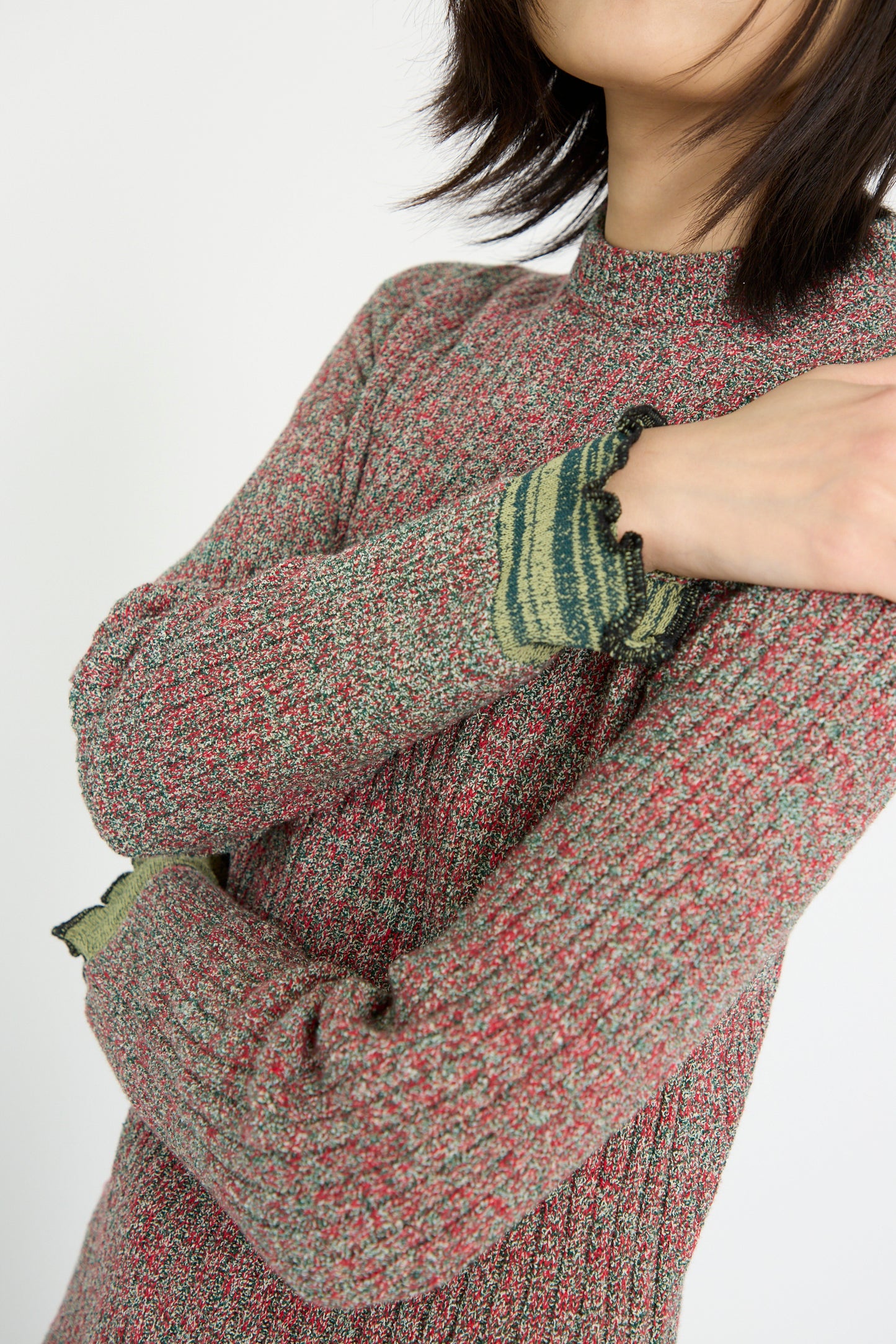 A person wearing the TOGA ARCHIVES Tweed Knit Back Zip Pullover in green, featuring speckled texture, green-trimmed cuffs, and a fringe hem, stands with crossed arms against a plain background.