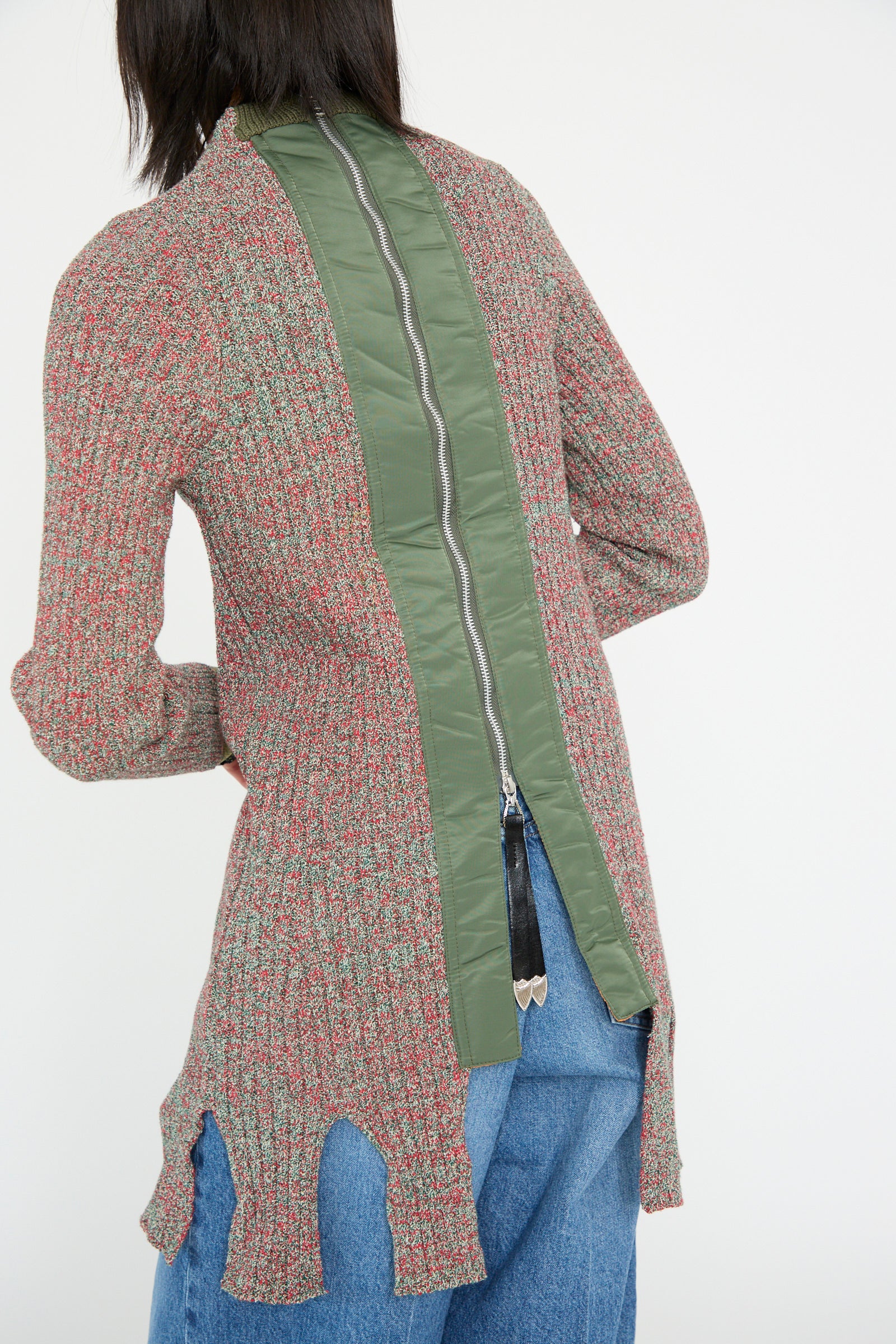 A person models the Tweed Knit Back Zip Pullover in Green by TOGA ARCHIVES, featuring a textured design with a fringe hem and central green zipper, paired with blue jeans and viewed from the back.