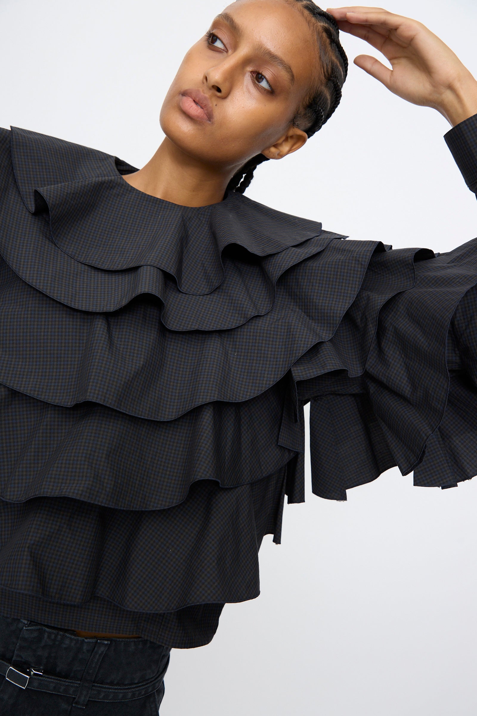 A person poses with one arm raised against a plain background, wearing the TOGA ARCHIVES Typewriter Check Frill Blouse in Navy, featuring ruffled layers, a boxy fit, and wide long sleeves.
