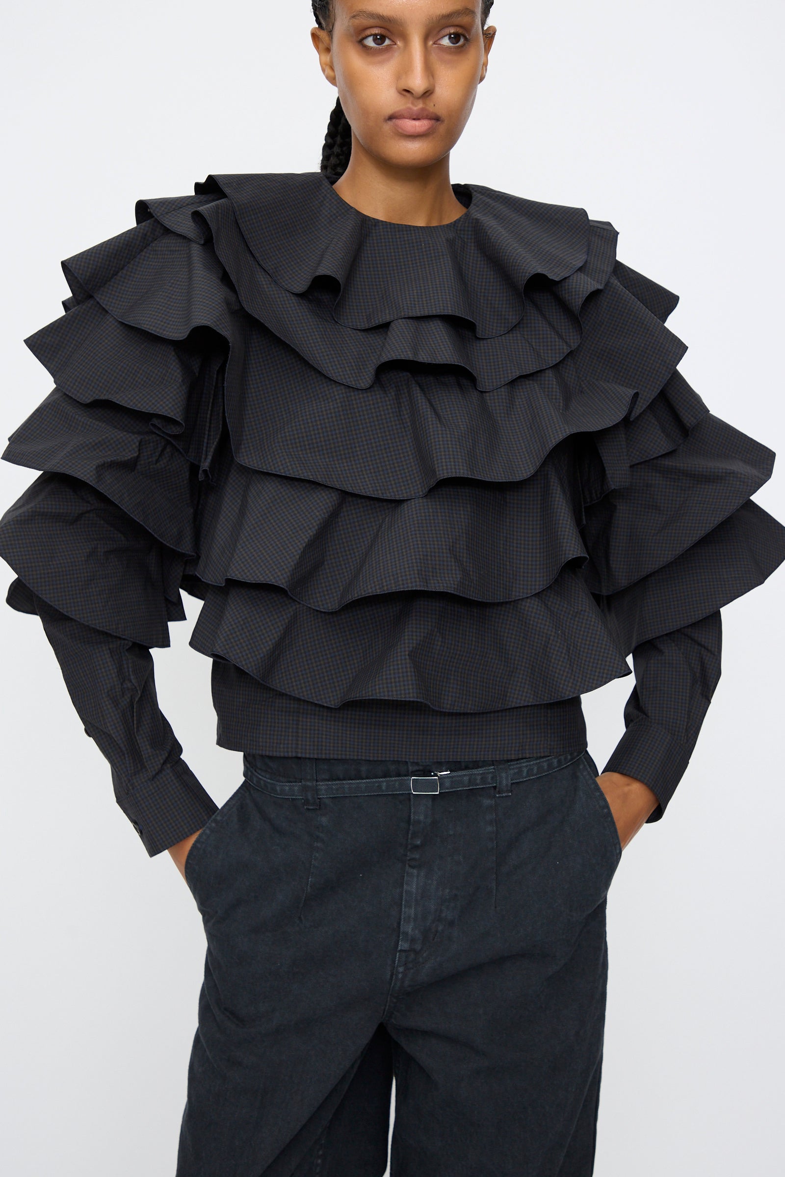 A person wears the TOGA ARCHIVES Typewriter Check Frill Blouse in Navy, featuring boxy long sleeves and multiple tiered ruffles on the sleeves and chest, paired with high-waisted dark pants against a plain background.