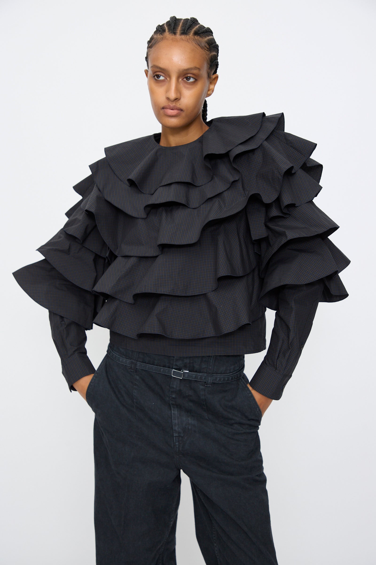 A person stands against a plain background wearing the TOGA ARCHIVES Typewriter Check Frill Blouse in Navy with layered ruffles and dark high-waisted pants, hands in pockets, looking forward.