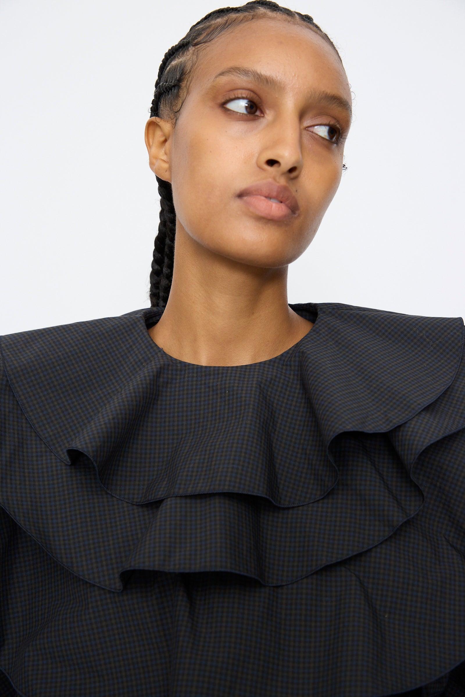 A woman with braided hair looks to the side, wearing the TOGA ARCHIVES Typewriter Check Frill Blouse in Navy, featuring large layered ruffles, against a plain light background.