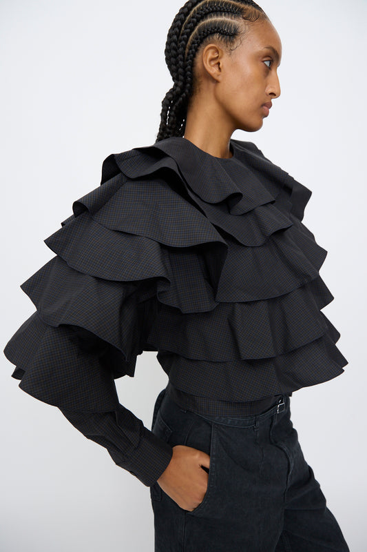 A person with braided hair, wearing the TOGA ARCHIVES Typewriter Check Frill Blouse in Navy and dark pants, stands in profile against a plain background.