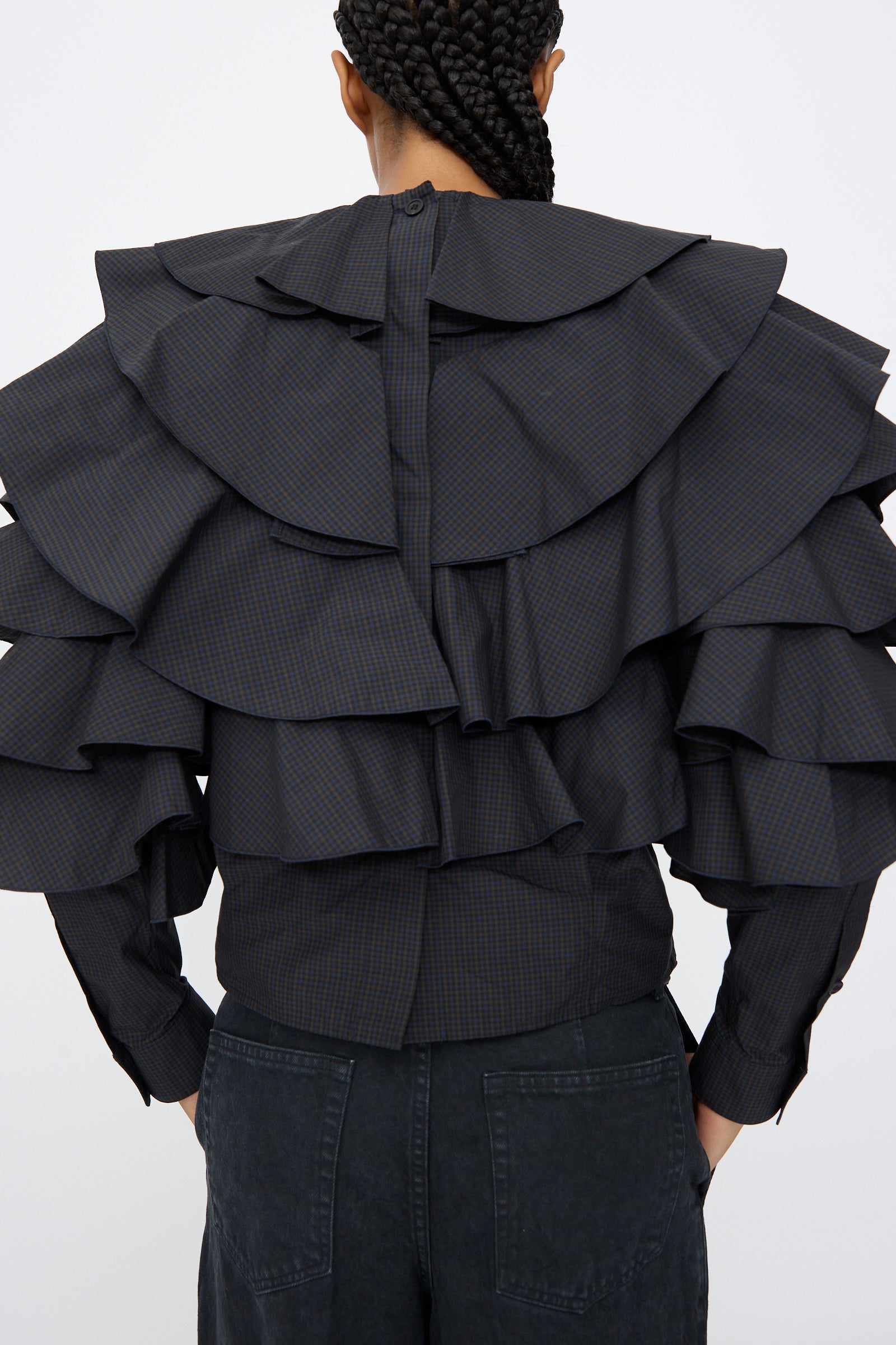 A person with braided hair is seen from behind wearing the TOGA ARCHIVES Typewriter Check Frill Blouse in Navy, featuring layered ruffles and a back zipper, paired with black pants.
