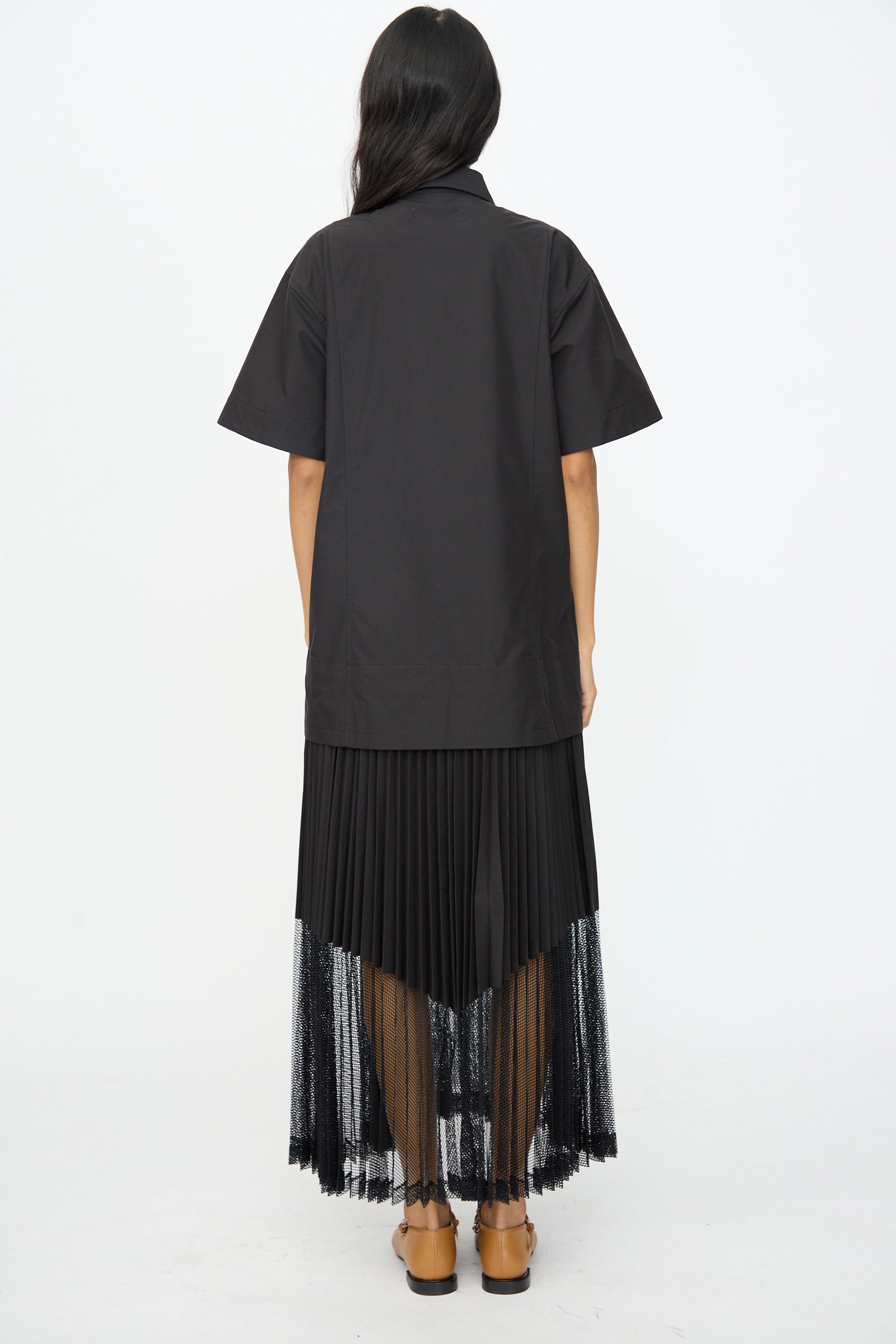 A person faces away, wearing the TOGA ARCHIVES Typewriter Mesh Dress in Black, featuring short sleeves and pleated details, paired with tan flat shoes.